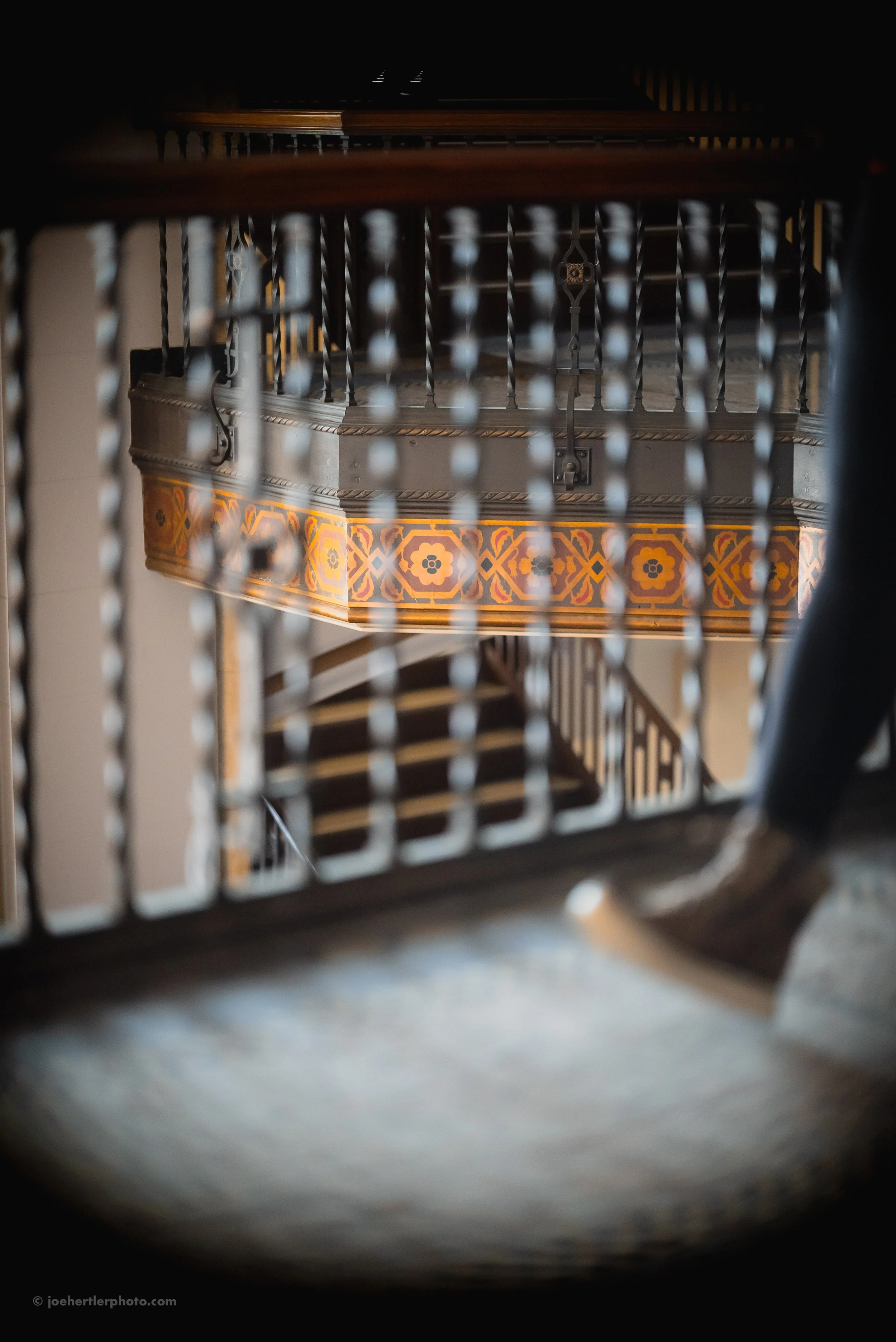 View of an ornate balcony or staircase railing through a blurred grid, with a staircase and decorative painted patterns in the background.
