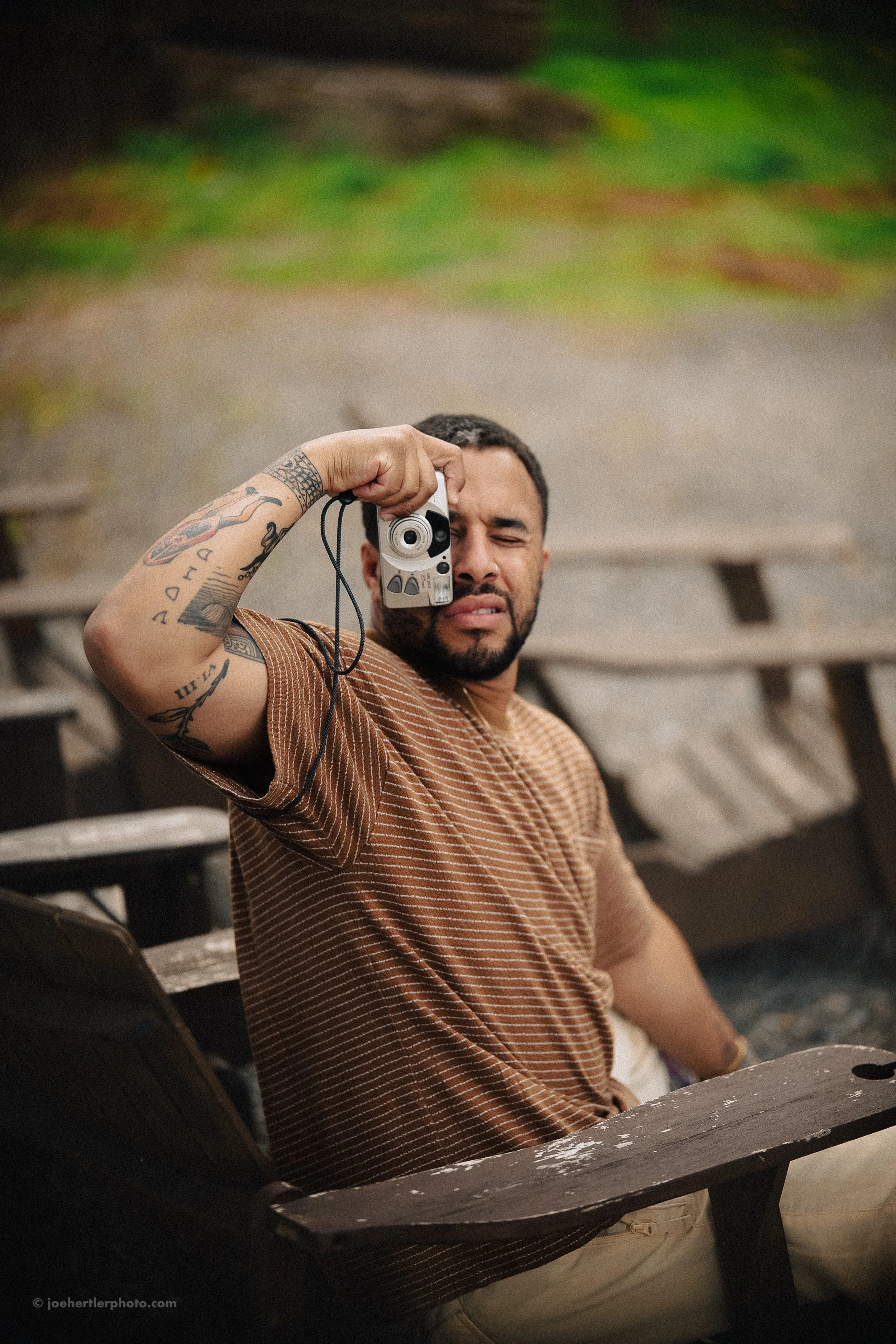 A man with tattoos, wearing a brown striped shirt, sitting outdoors on a wooden bench, taking a selfie with a vintage film camera. He is squinting or grimacing and behind him is a blurred background of green foliage.