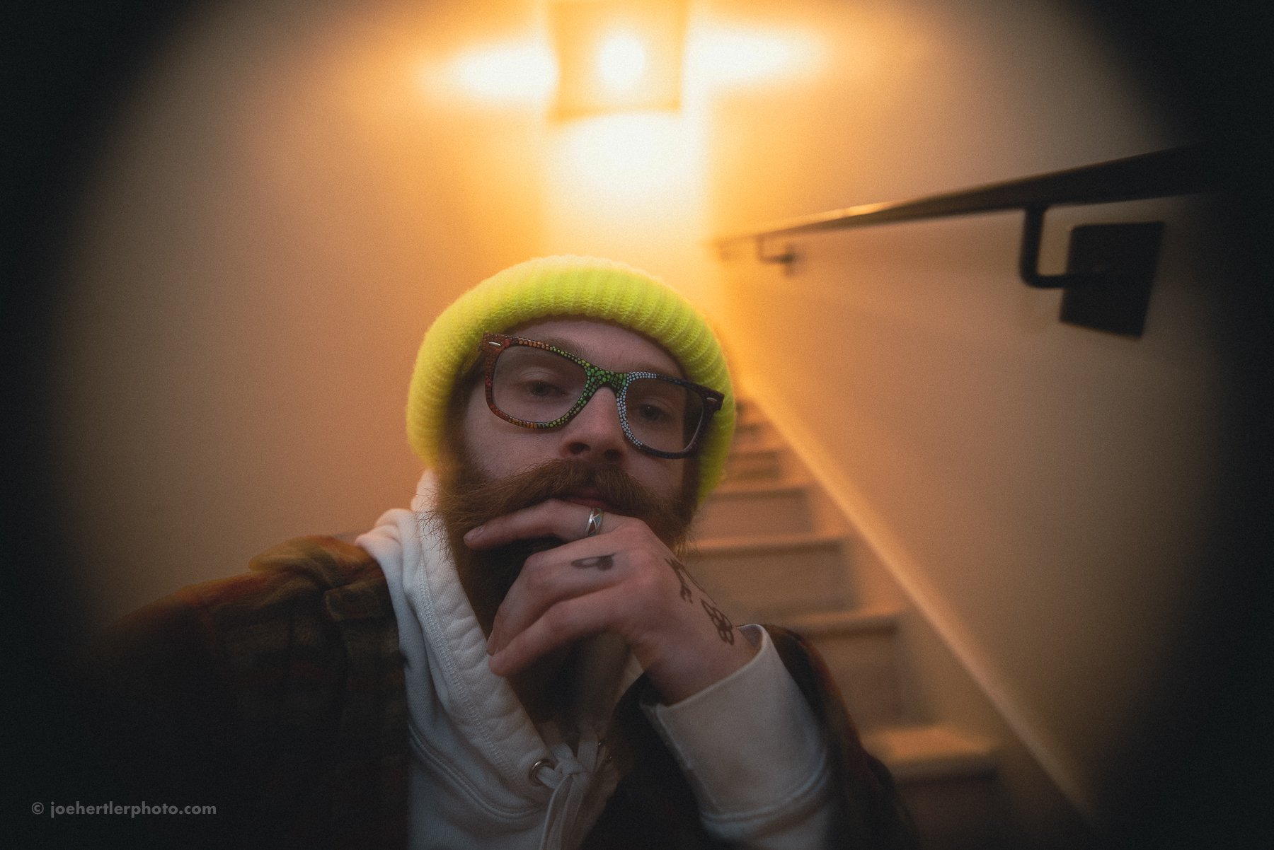 A man with a beard, glasses with green and red rhinestones, yellow beanie, and plaid jacket, photographed from below near a staircase with warm lighting.
