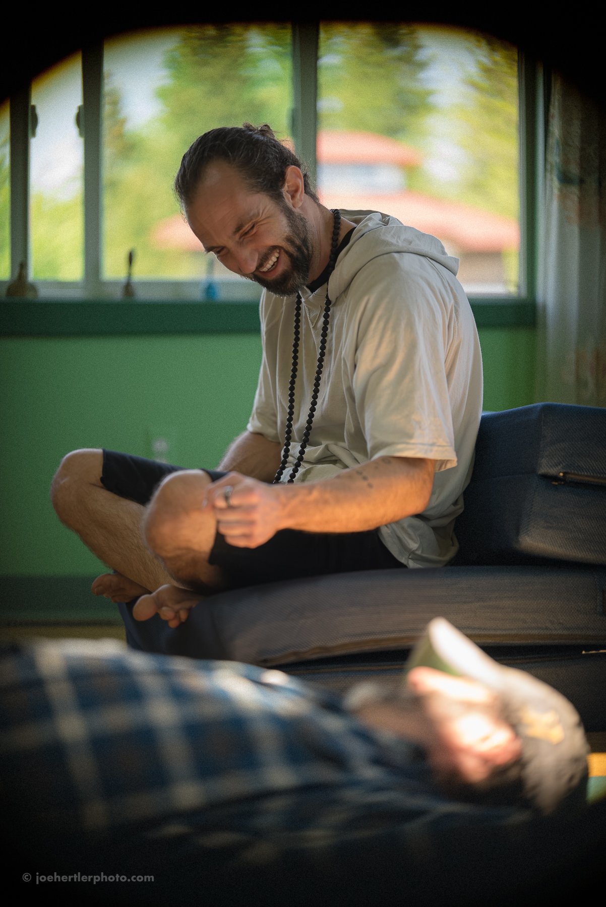 A man with long hair, a beard, wearing a light-colored hoodie and black shorts, is sitting on a bed, smiling and laughing while looking at someone lying on the bed. The person lying down is partially visible, wearing a plaid shirt and holding a cup. 