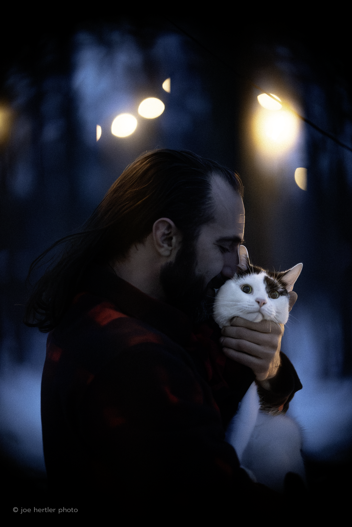 A man with long hair and a beard holding a white cat with black markings close to his face at night, with streetlights in the background.
