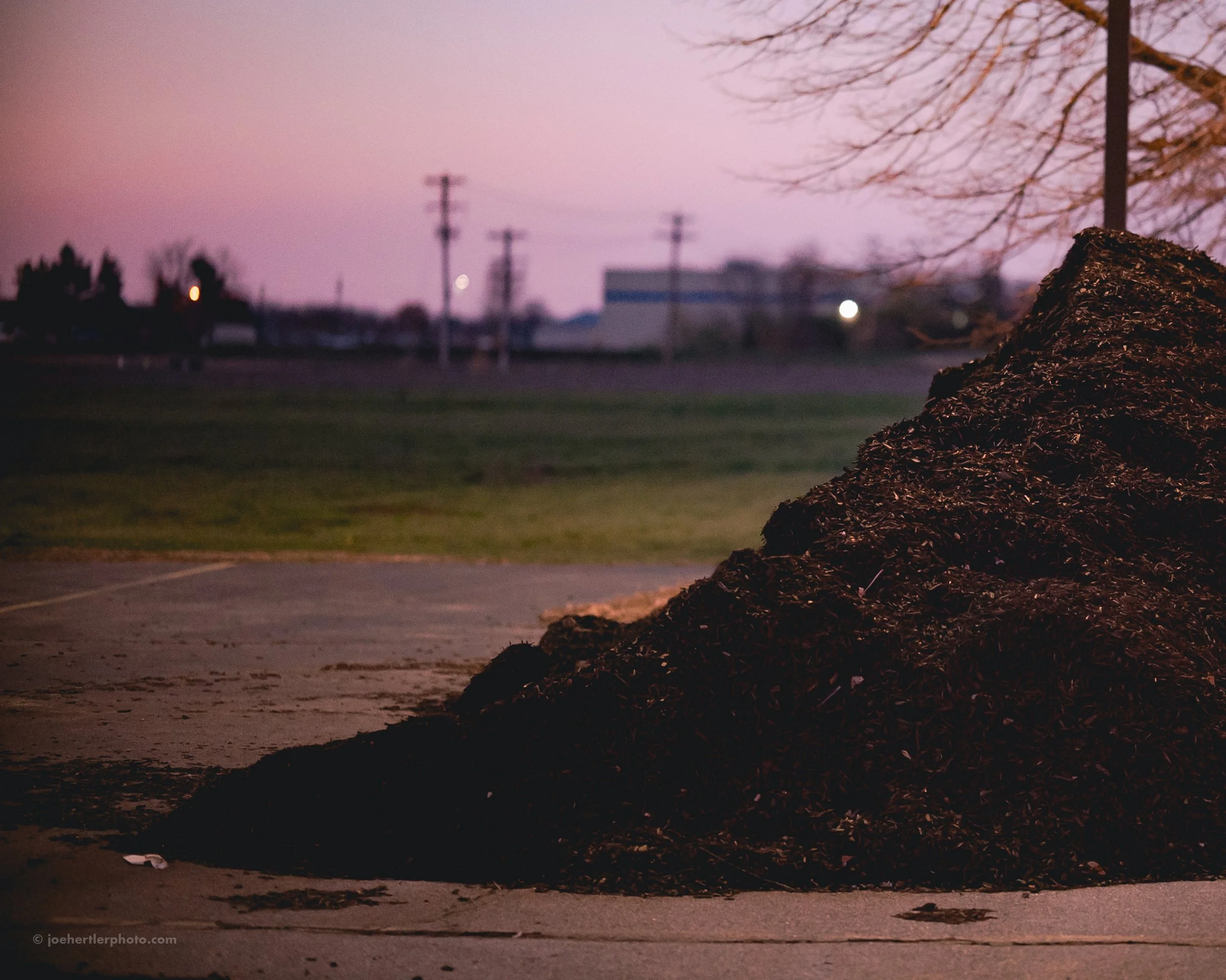 A large pile of dark mulch on a concrete surface outdoors during dusk, with a blurred background of utility poles, grass, and sparse trees under a pinkish-purple sky.