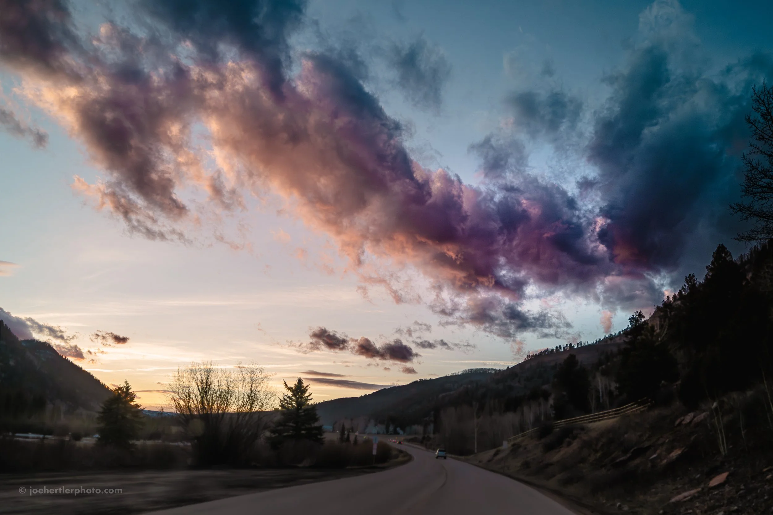 A winding mountain road at sunset with a sky filled with dark storm clouds and hints of pink and orange