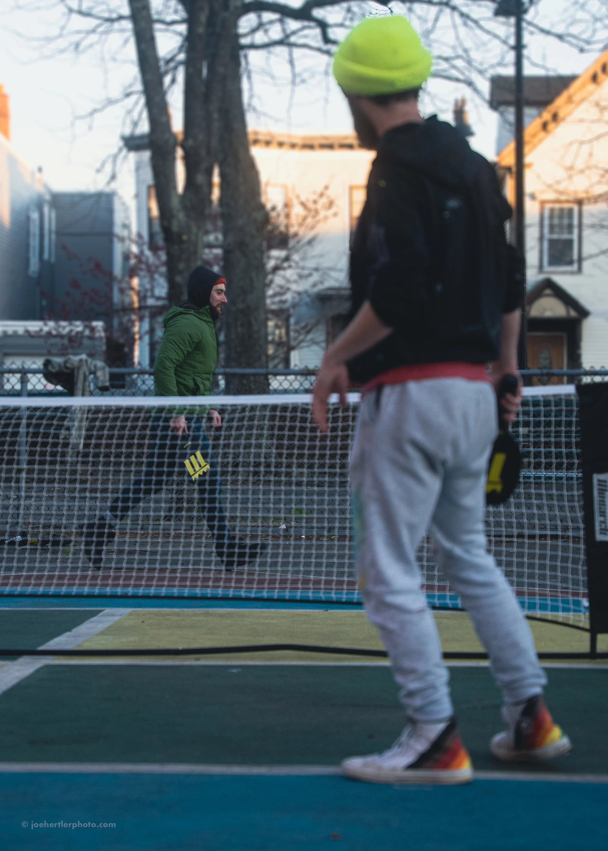 Two men playing pickleball outdoors on a court with trees and houses in the background. One man, in a black shirt and yellow beanie, is facing away from the camera, while the other man, in a green jacket, is behind a net.
