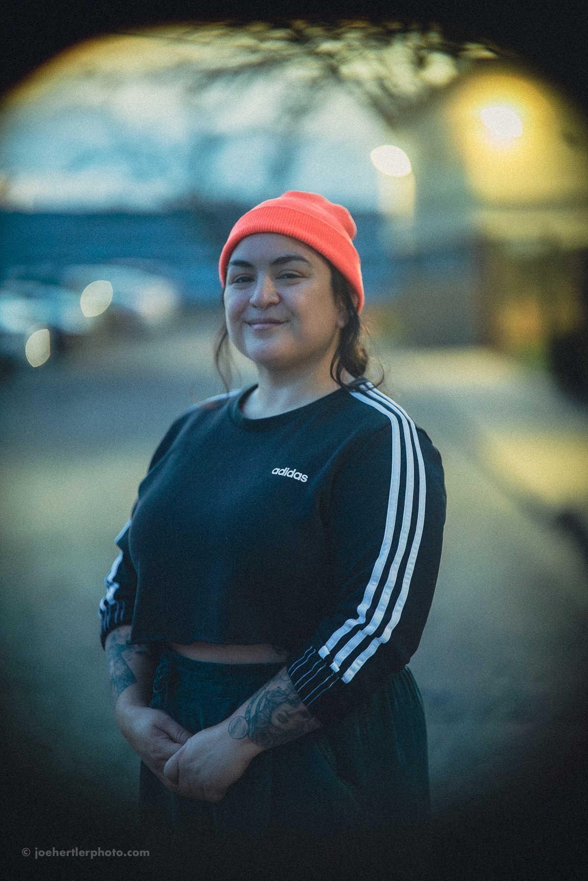 A woman wearing a red beanie and a black Adidas top with white stripes on sleeves, standing outdoors during sunset, smiling softly at the camera.