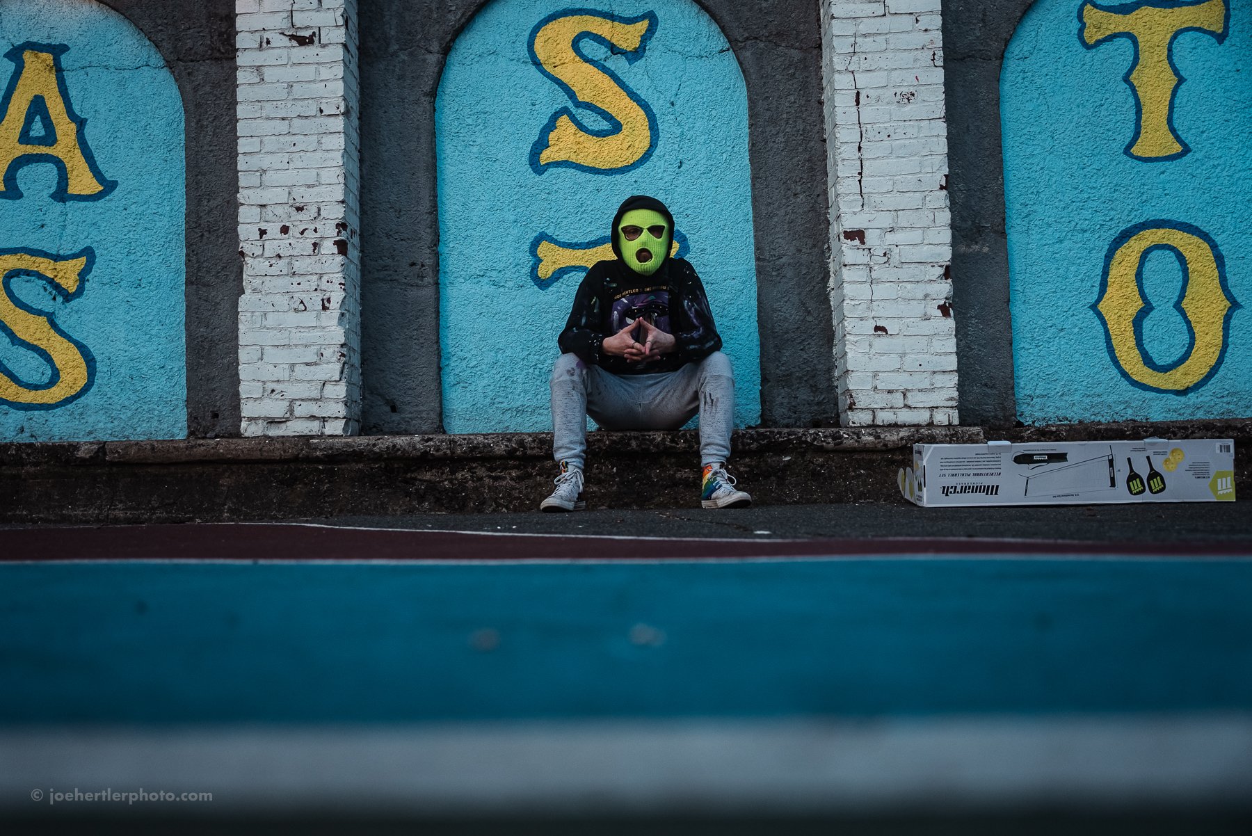 A person wearing a green mask, sunglasses, and a black jacket sits on the ground against a graffiti-covered wall, with a boxed BBQ grill nearby.
