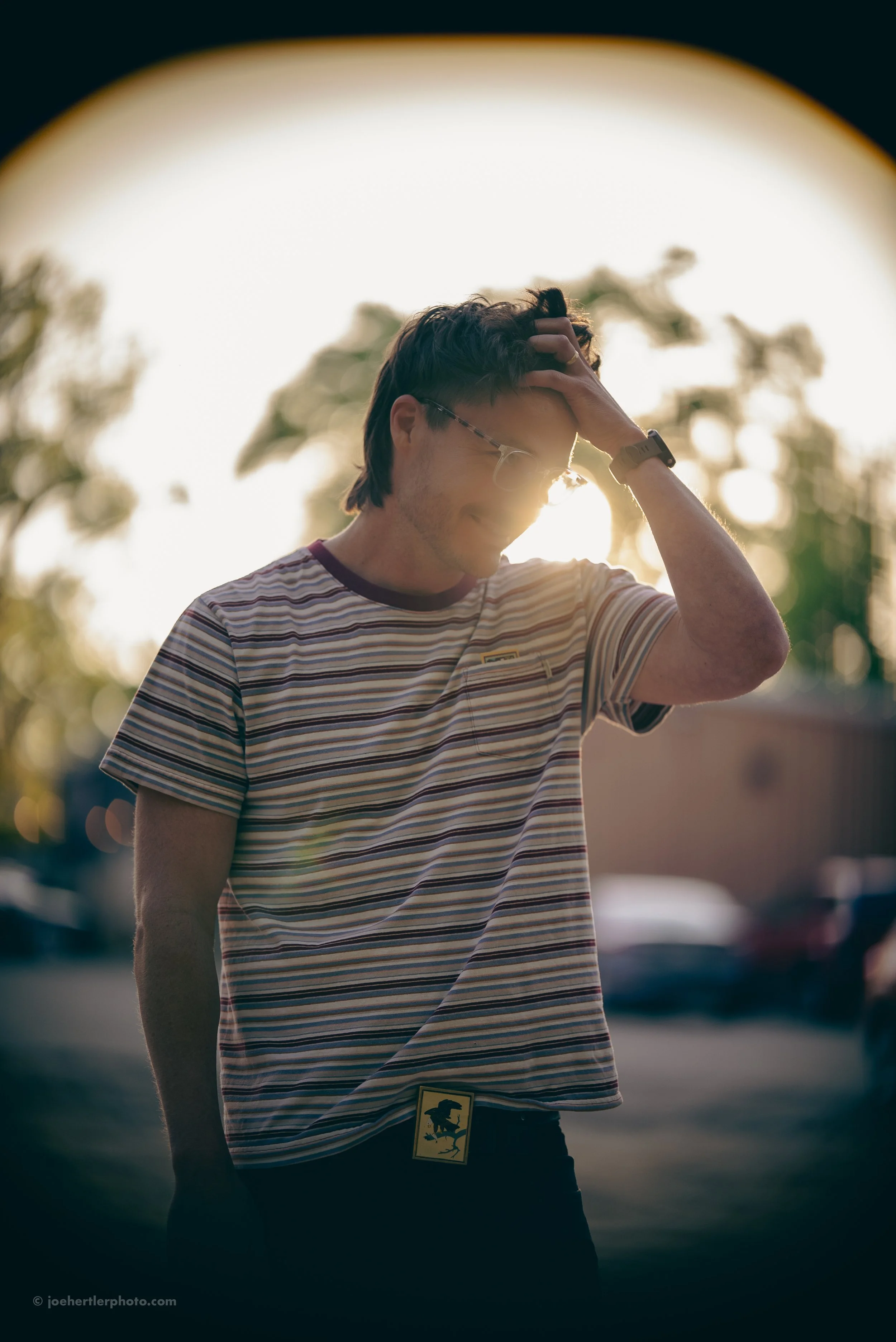 A young man with glasses and a striped shirt smiling with his hand on his head, standing outdoors near sunset.