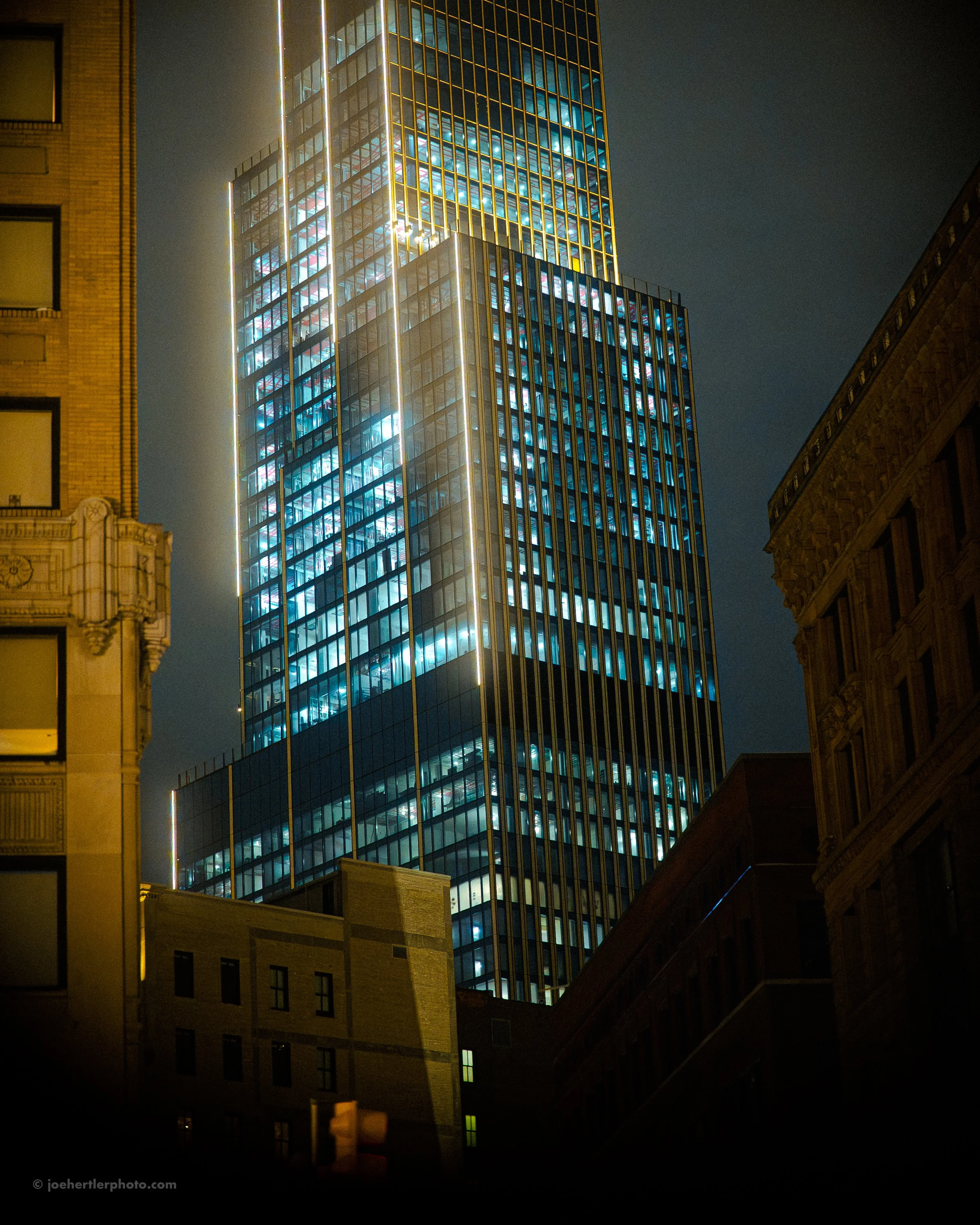 Night view of a tall modern glass skyscraper with illuminated windows in a city, surrounded by older brick buildings