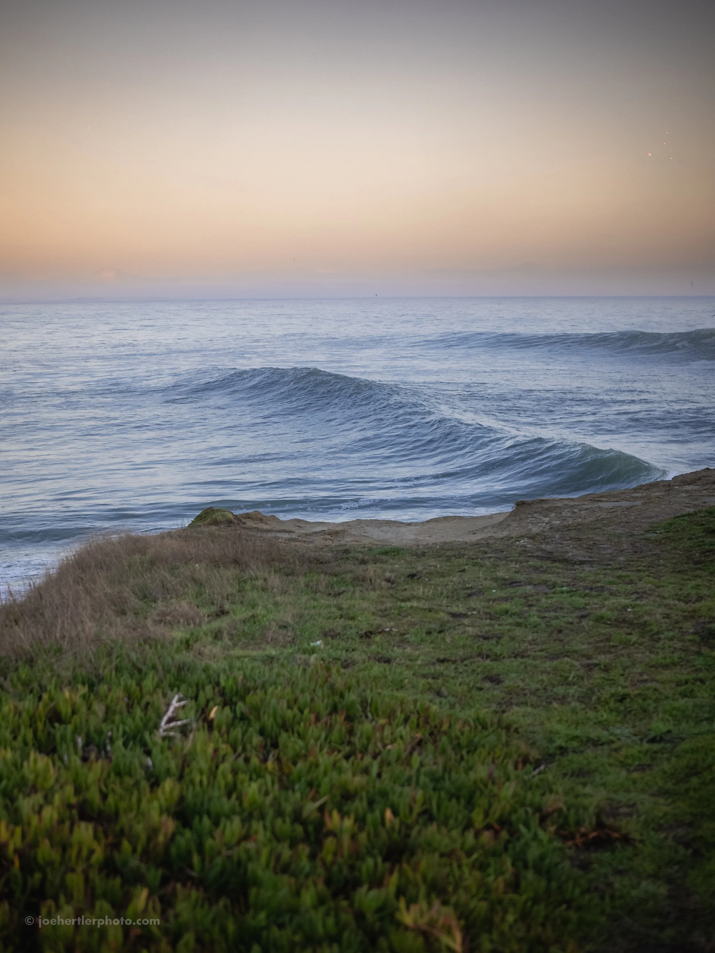 View of the ocean with gentle waves, partly cloudy sky at sunset, and green grassy foreground.