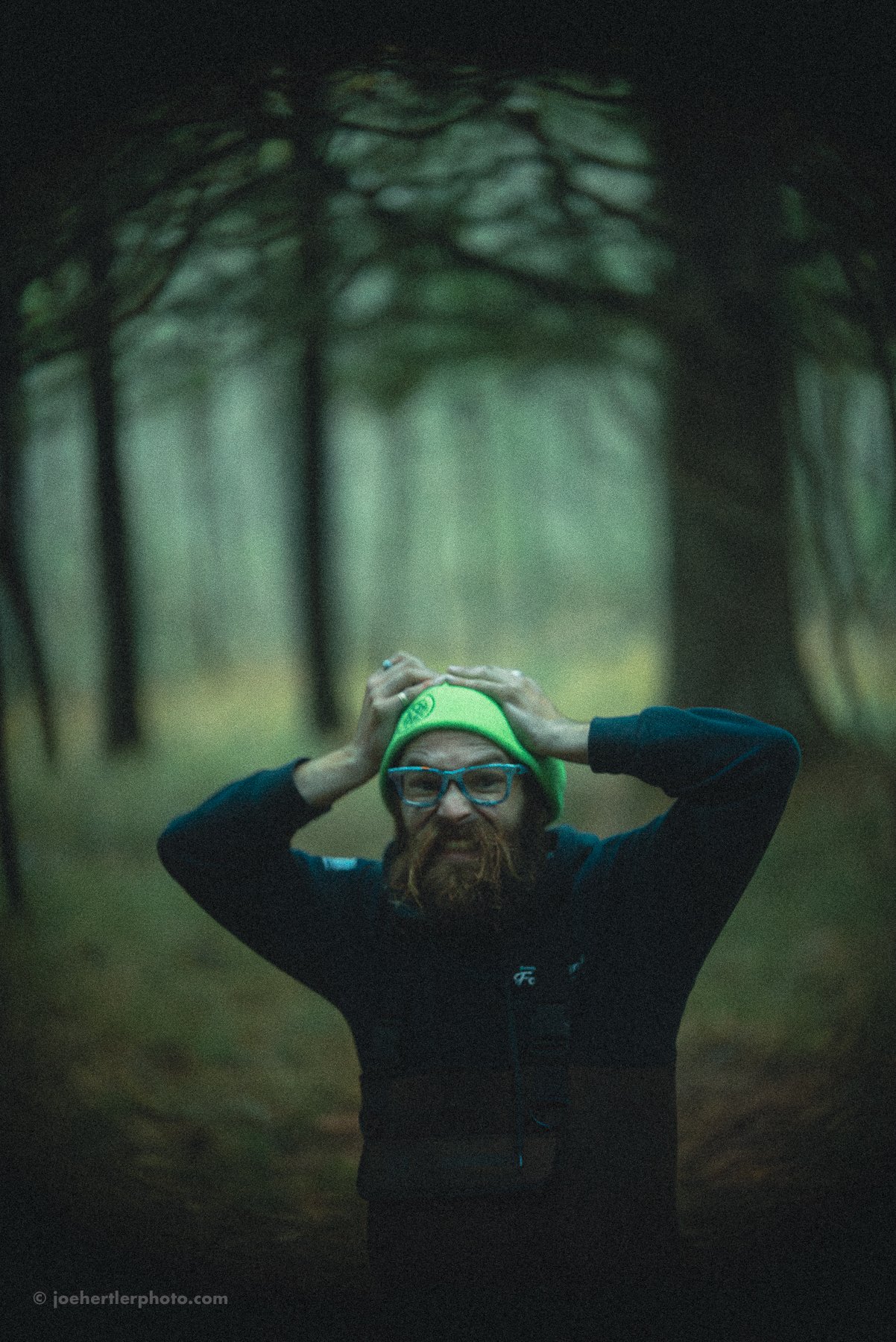A bearded man with glasses and a neon green beanie, standing on a forest trail, clutching his head with both hands, with tall trees surrounding him.