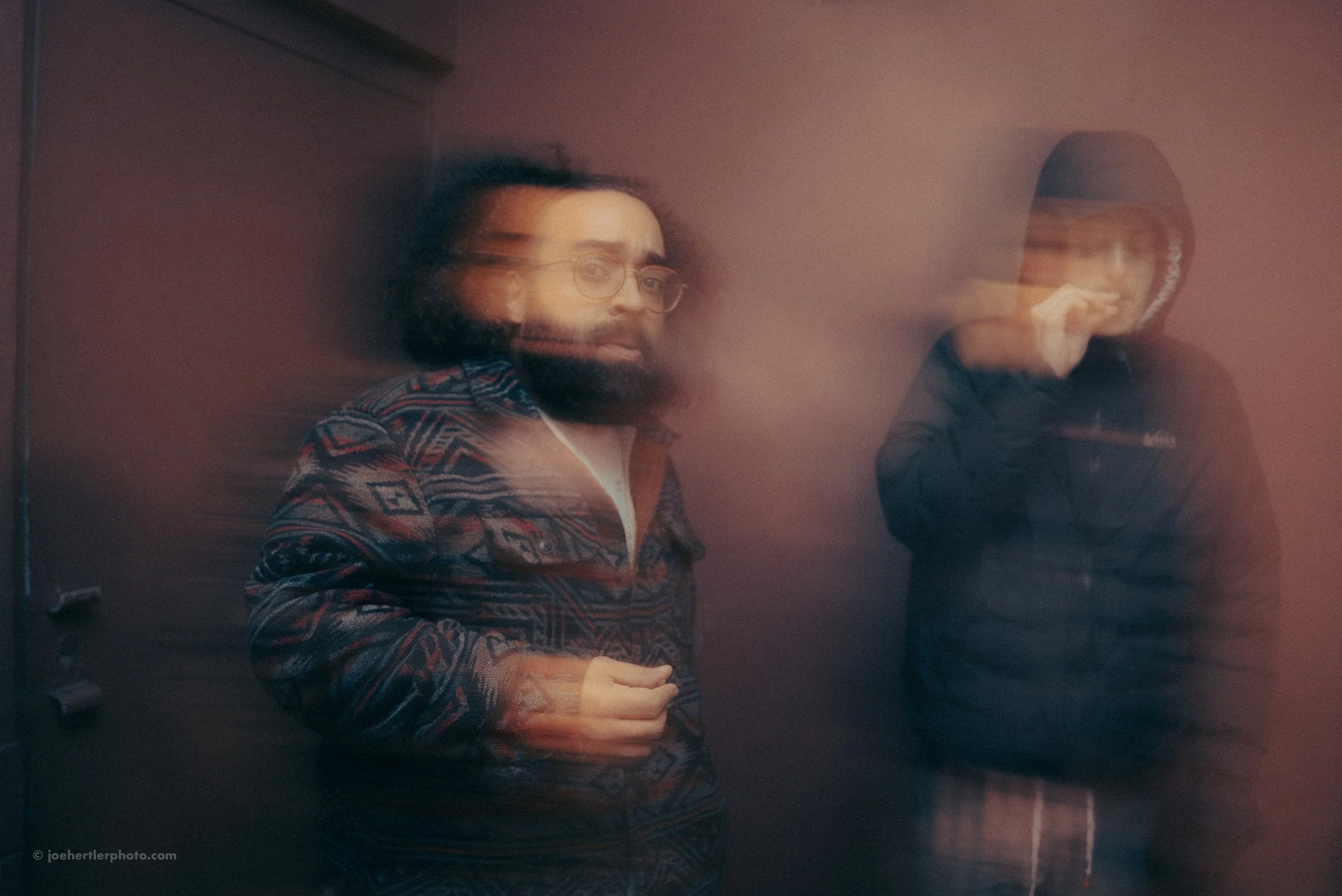 Two young men standing against a wall, with a motion blur effect creating a ghostly appearance. The man on the left has glasses, a beard, and is wearing a patterned jacket. The man on the right has dark hair and is wearing a dark hoodie and jacket, h