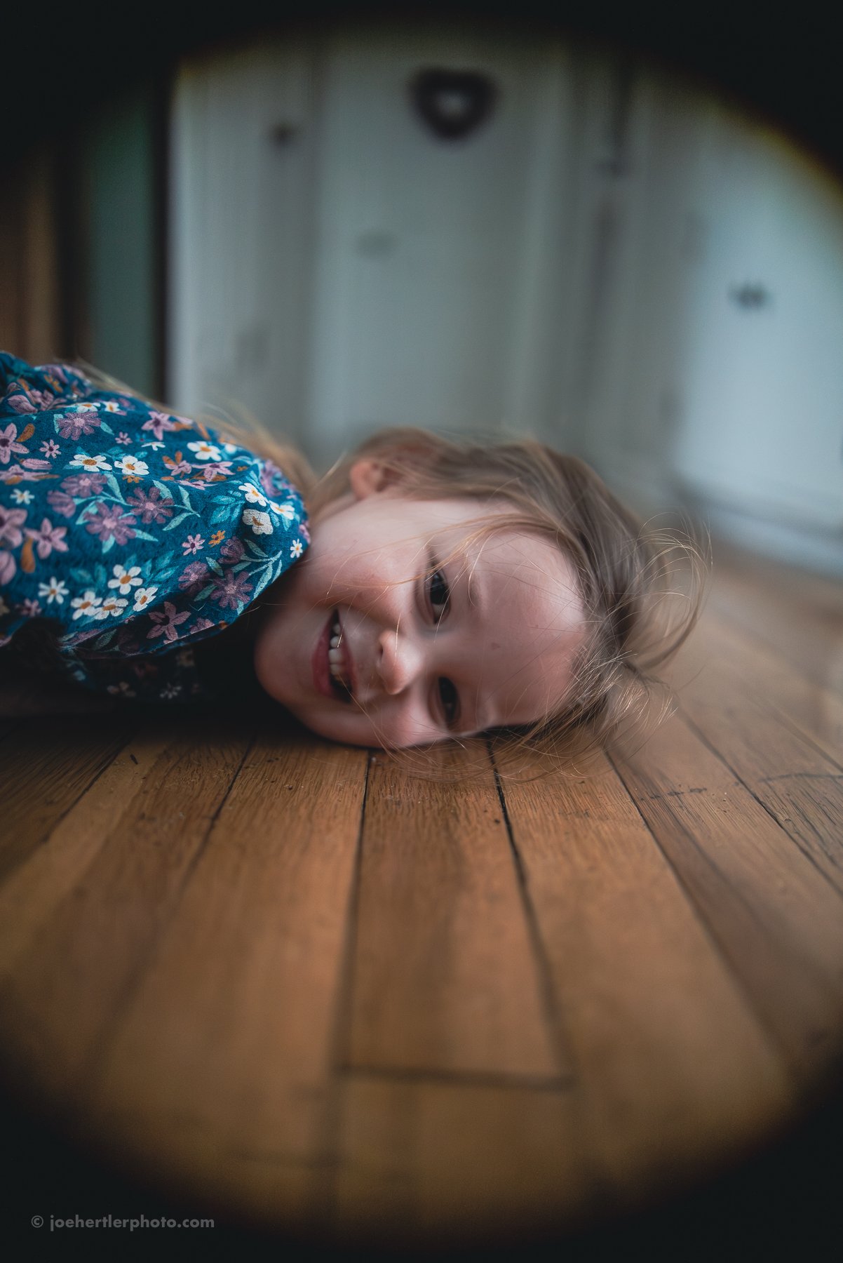 A young girl lying on a wooden floor, smiling and looking into the camera, with a blurred background.