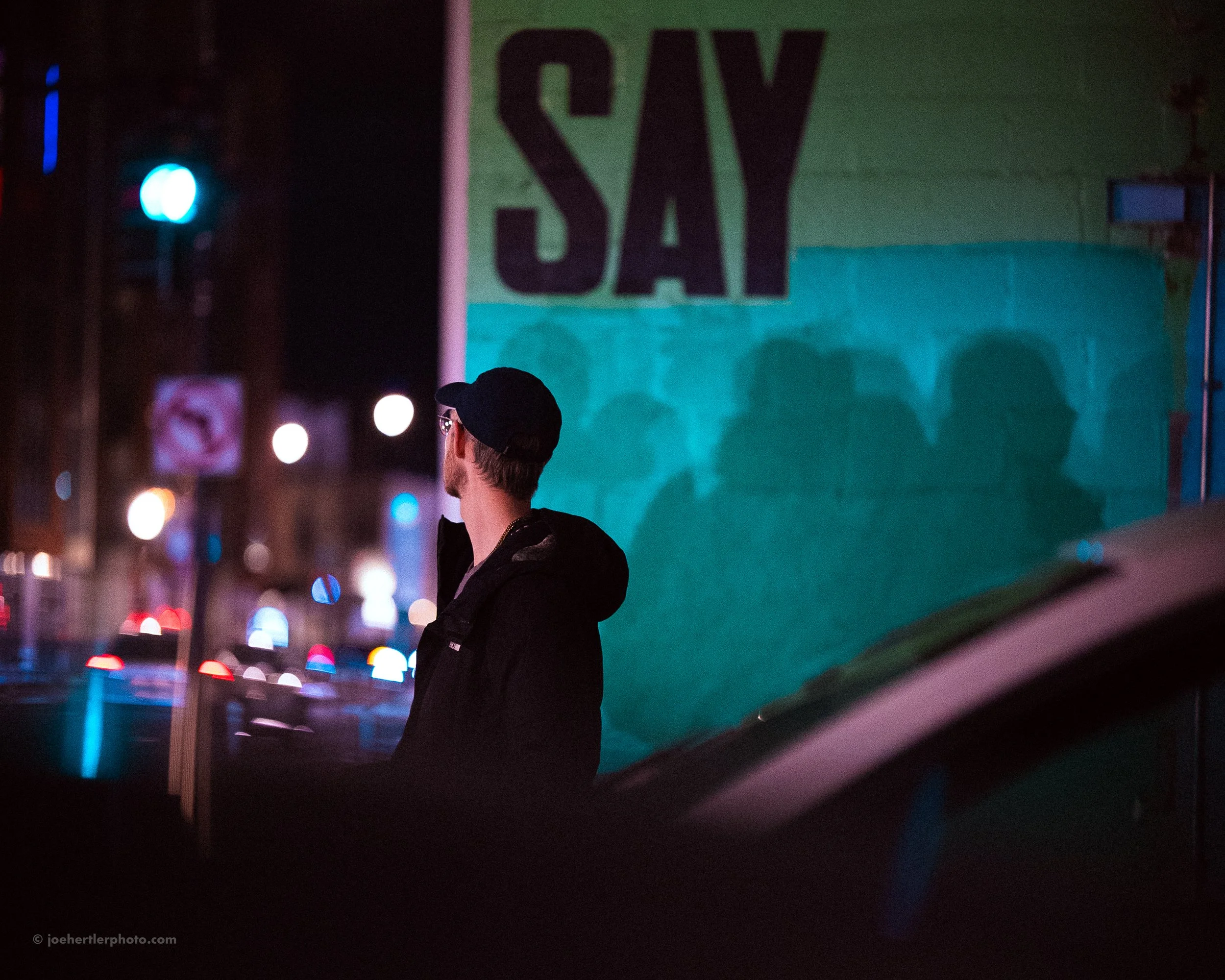 A man in a dark jacket and baseball cap stands on a city street at night, looking away from the camera, with a mural of the word 'SAY' and shadows of people cast on it in the background.