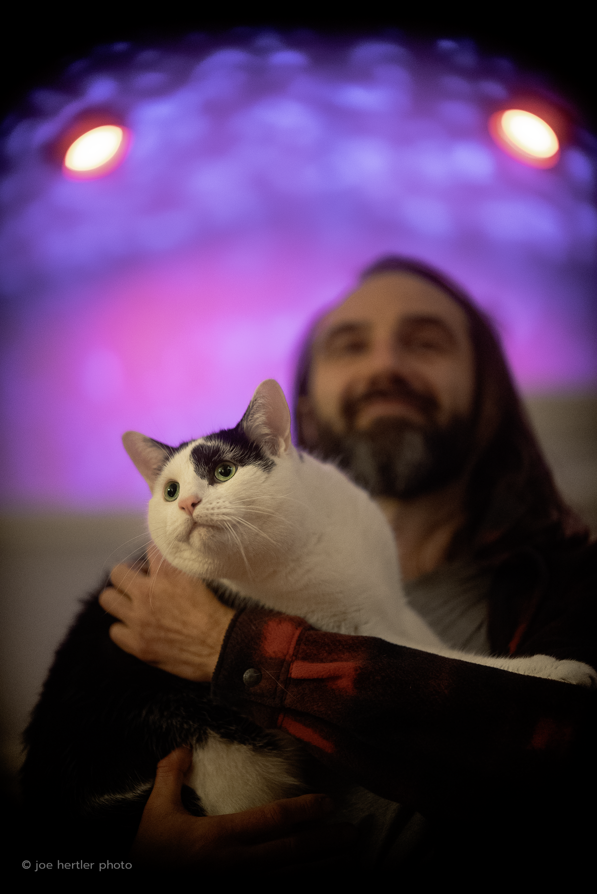 A man with long hair and beard holding a black and white cat in front of a colorful, glowing background.