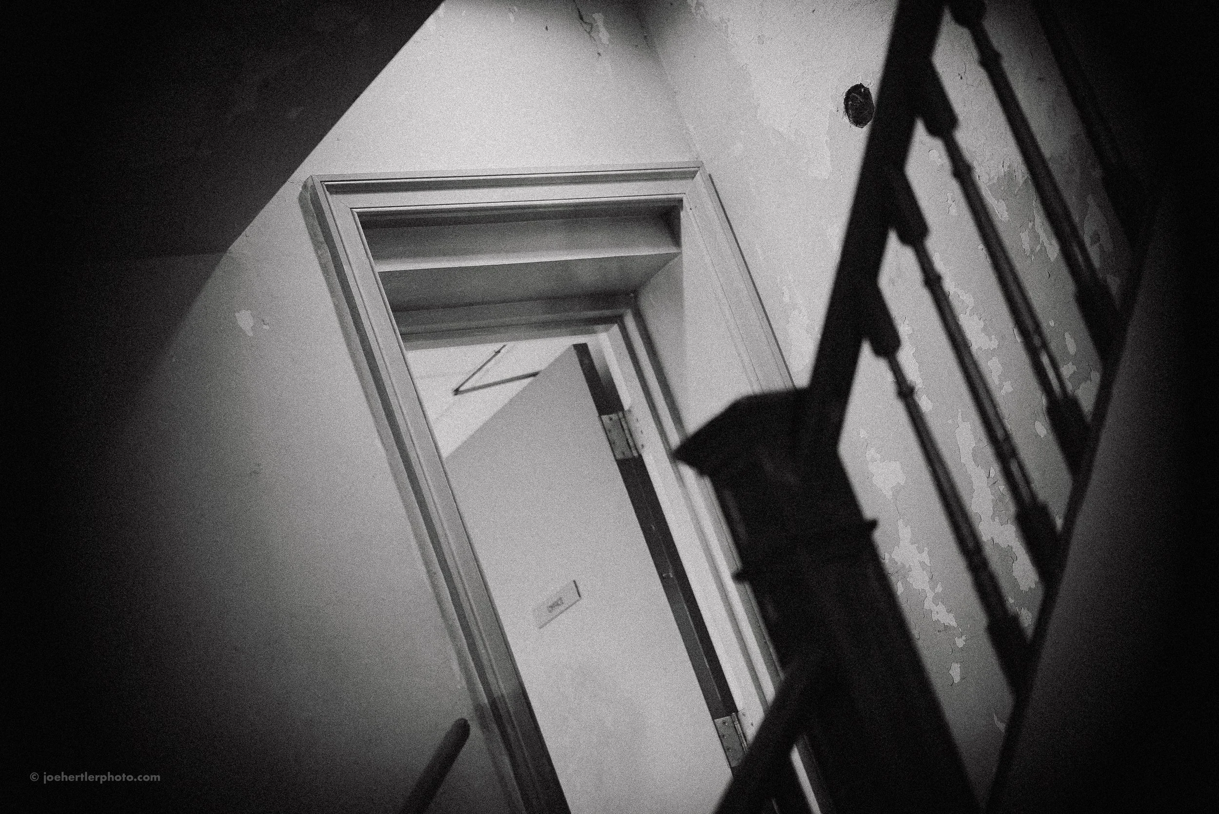 Black and white photo of an attic space with a doorway leading to another room. A ladder is leaning against the wall in the foreground, partially obscuring the view.