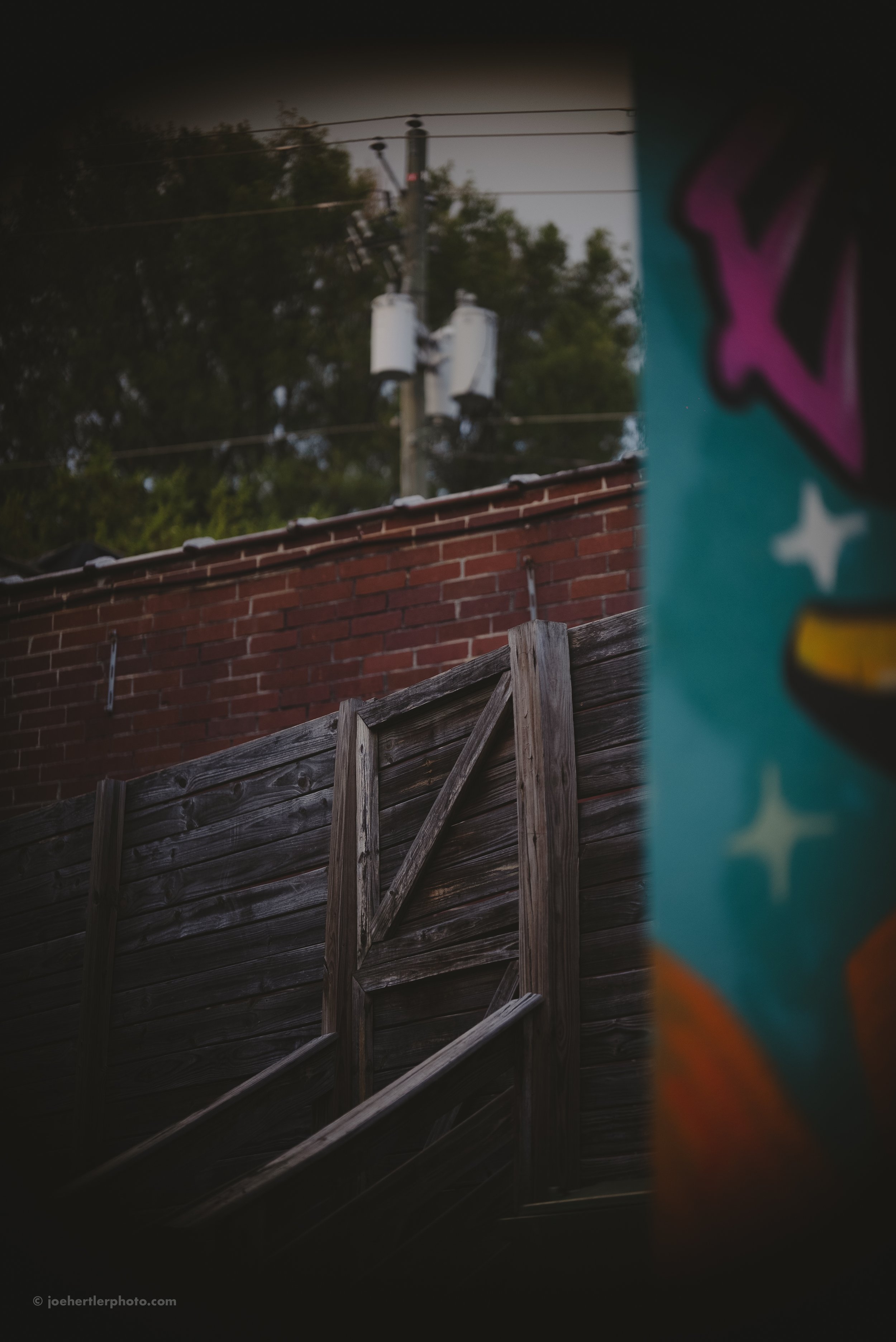 View through a small hole showing a wooden fence, a brick wall, and an electric pole with transformers in the background, partially obscured by a colorful graffiti wall on the right.