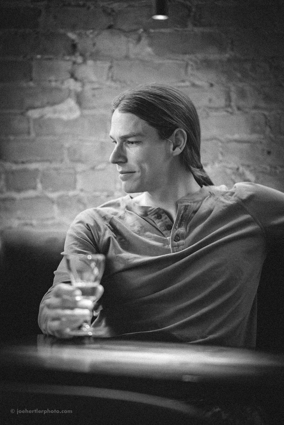 Black and white photo of a man with long hair in a ponytail, sitting at a table with a glass of water in his hand, in front of a brick wall.