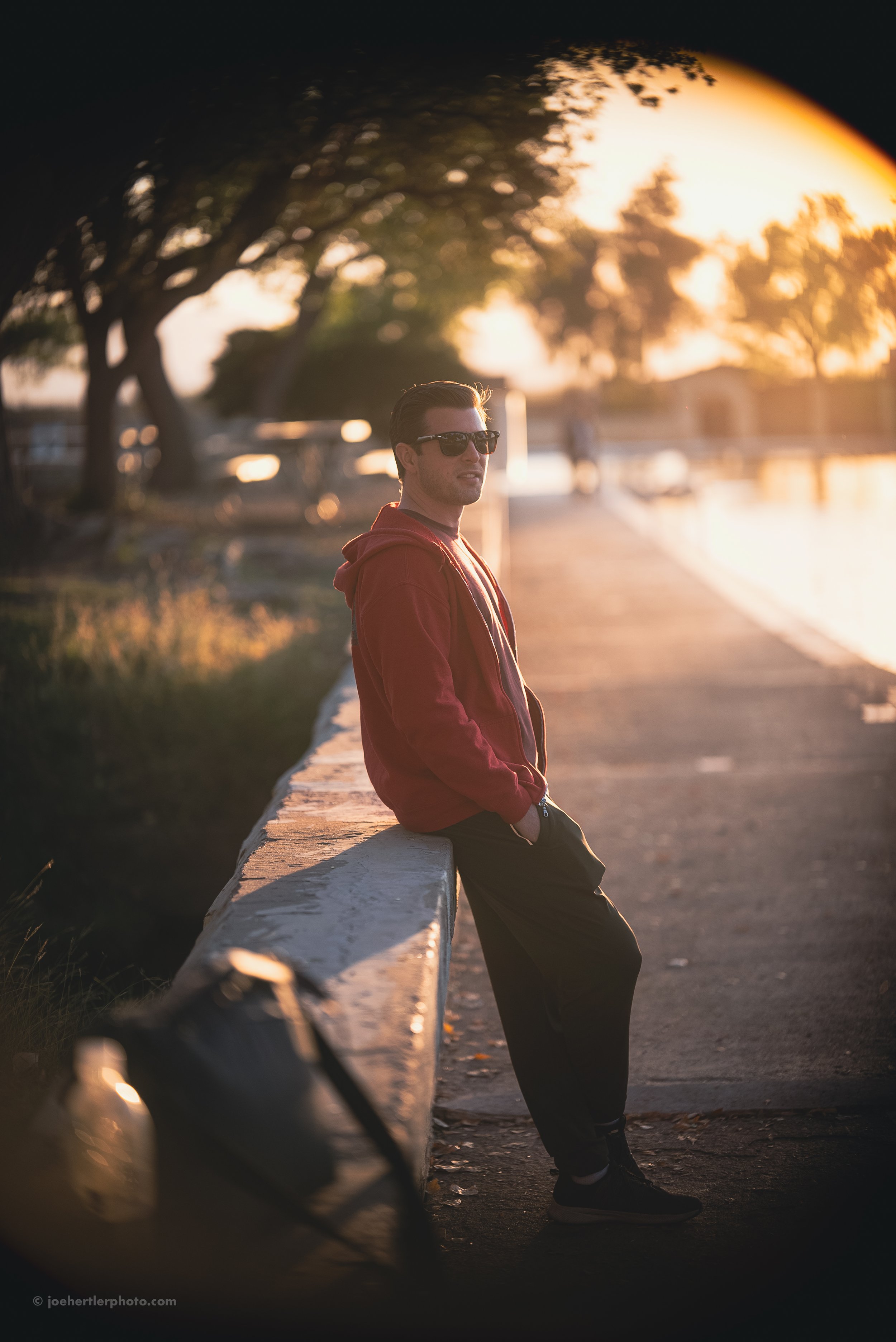 A man wearing sunglasses and a red hoodie lean against a concrete railing along a waterfront at sunset, with trees and a walking path in the background.