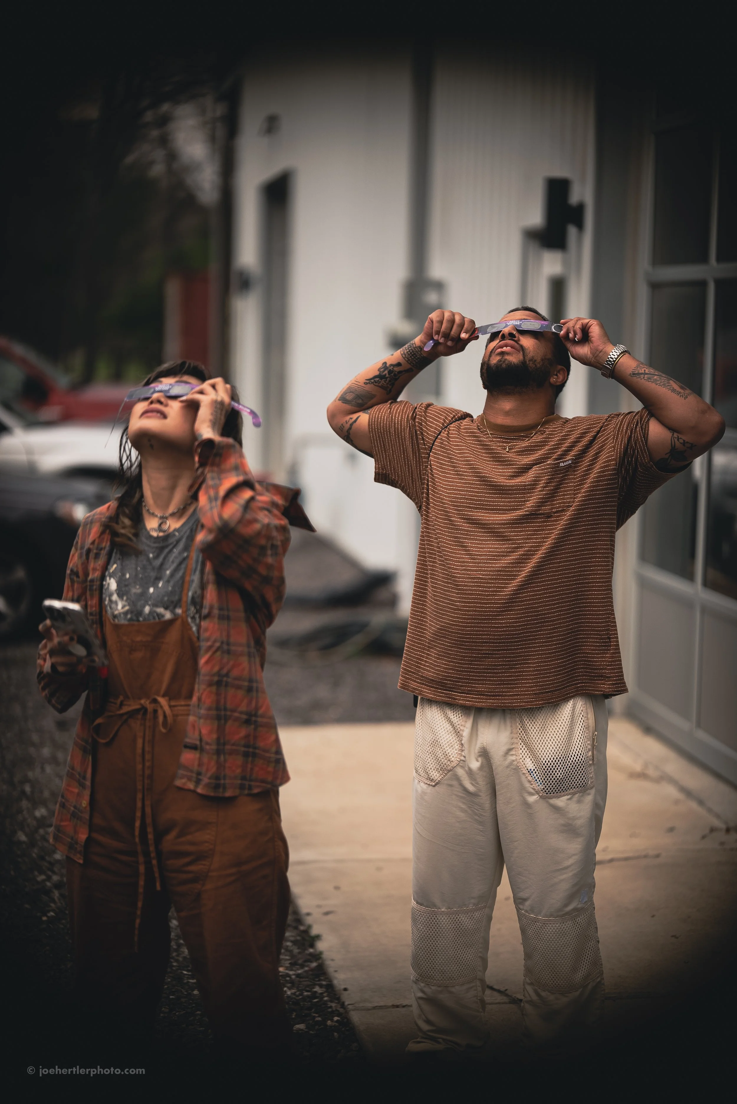 Two people standing outside looking upward while holding protective glasses, one woman with long dark hair wearing a plaid shirt and overalls, and one man with tattoos wearing a brown striped shirt and light-colored pants.