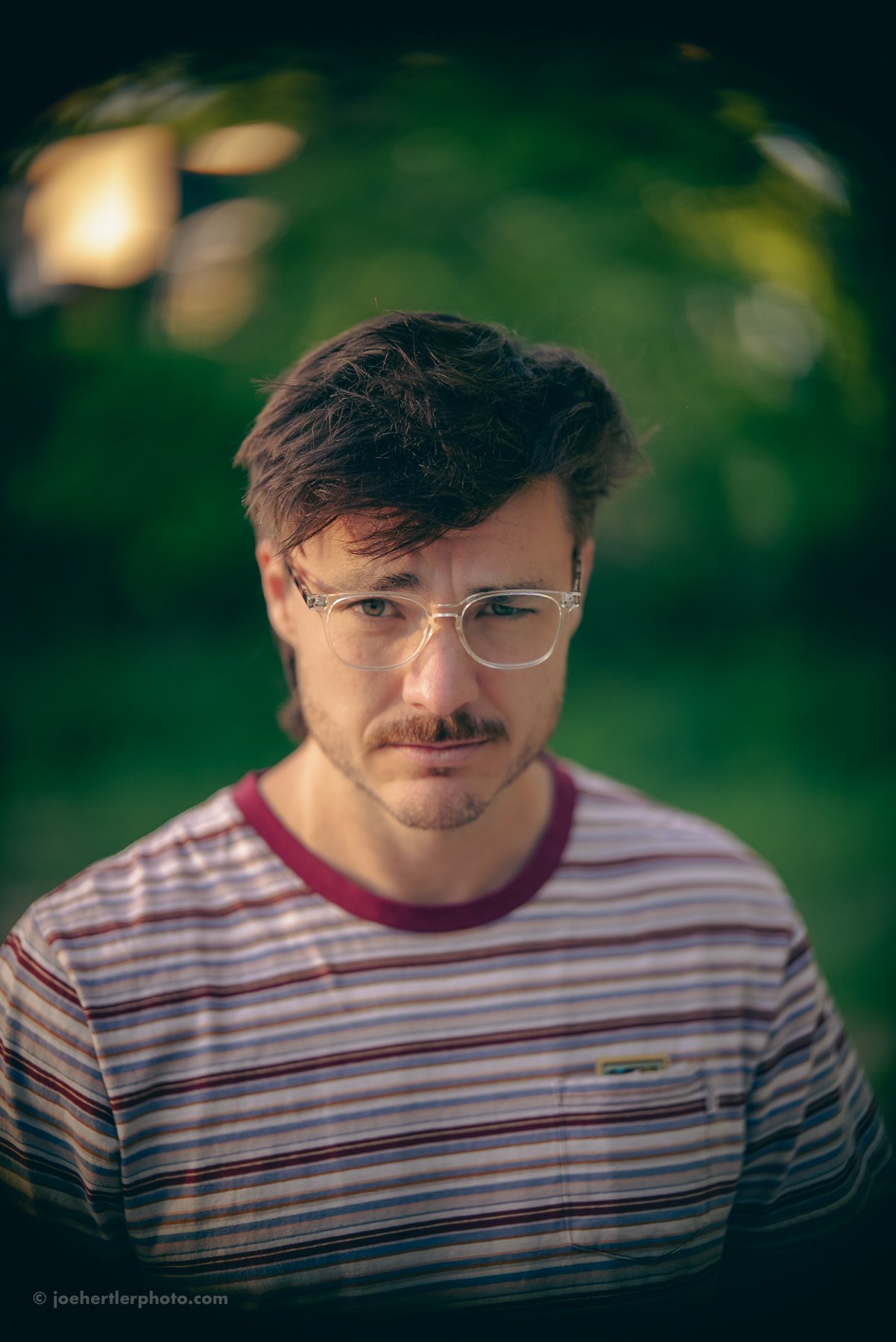 Serious man with glasses and a striped shirt outdoors with blurred green background.