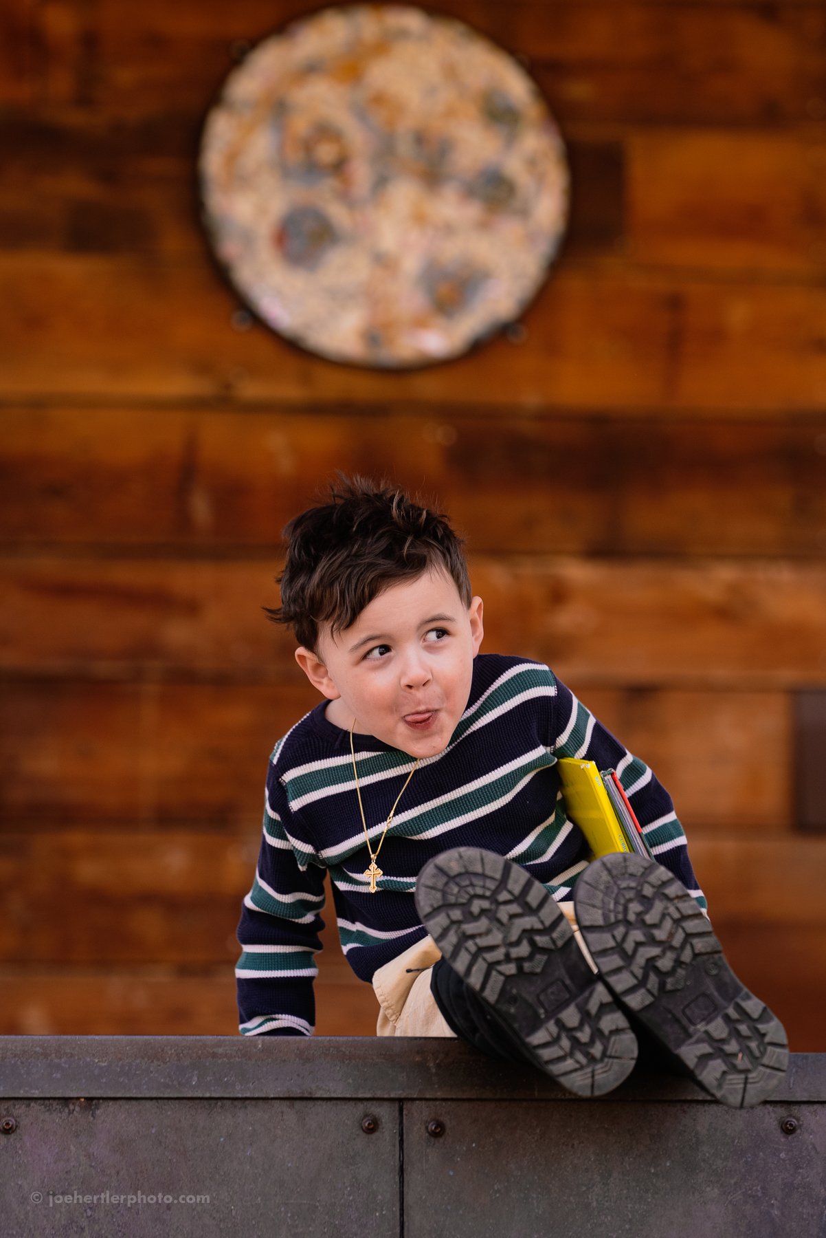 A young boy with dark hair, wearing a striped sweater, kneeling on a table with his right foot raised, sticking out his tongue and looking to the side. There is a wooden wall and a pizza on a wall hanging in the background.