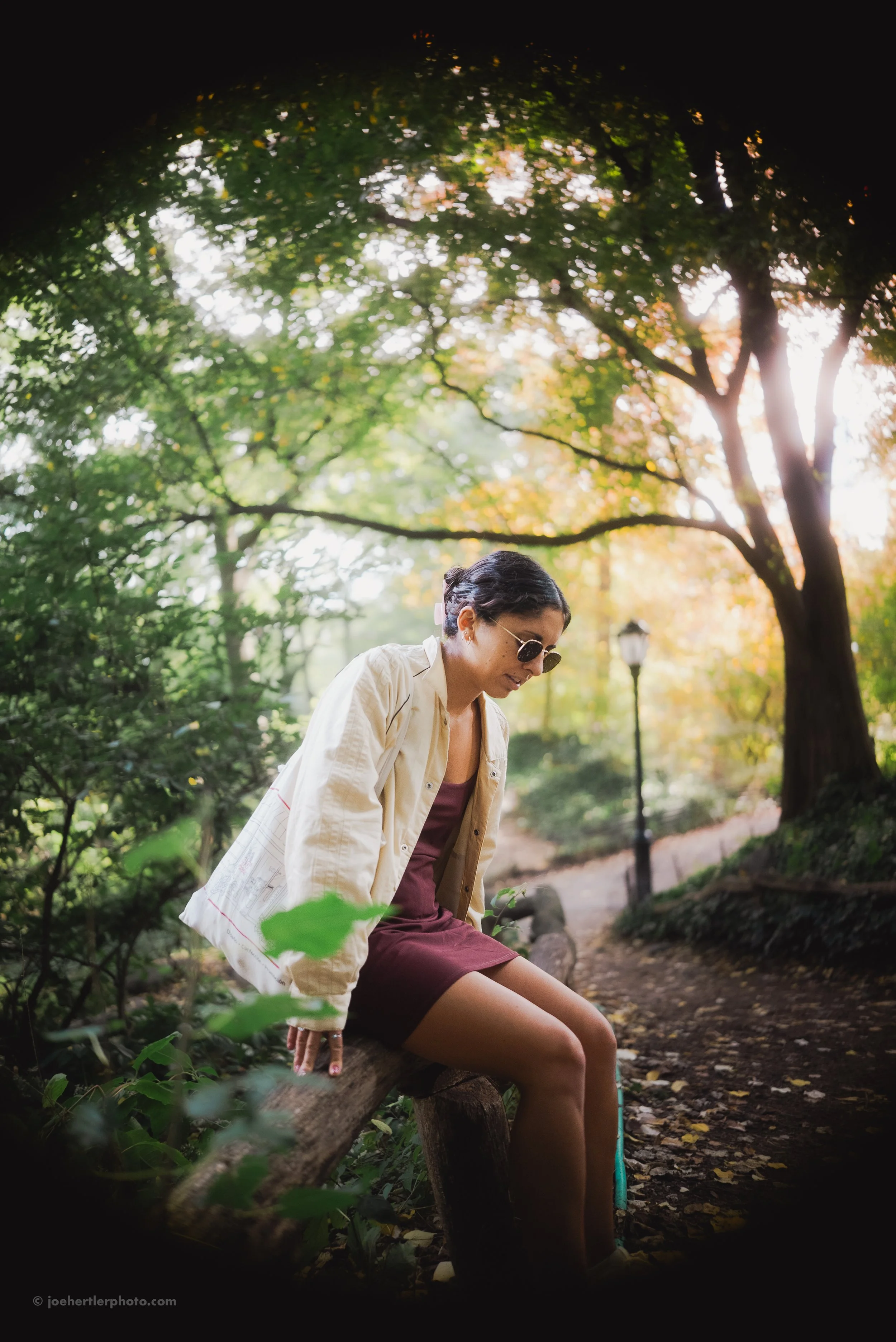 A woman sitting on a wooden bench in a park surrounded by trees with green and orange leaves, wearing sunglasses, a beige jacket, and a maroon dress, during what appears to be late afternoon or early evening sunlight.