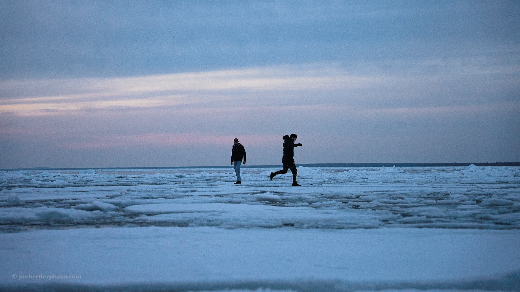 Two people walking on an icy, snow-covered landscape under a cloudy sky during sunset or sunrise.