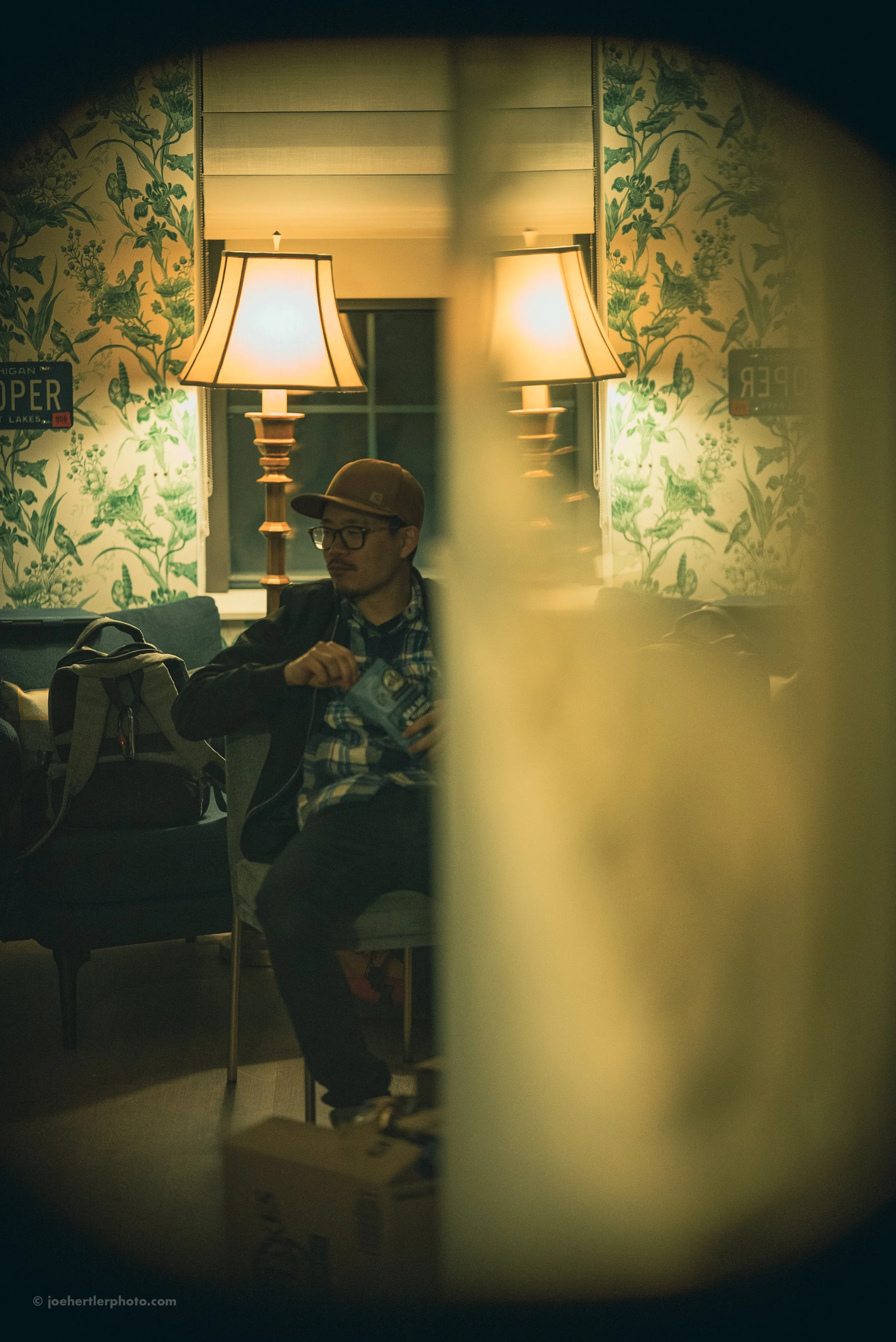 A man wearing glasses, a brown cap, and a black jacket sitting on a chair in a dimly lit room, holding a snack bag, with a backpack on a sofa next to him. The room has green floral wallpaper, two table lamps, and a window with white blinds.