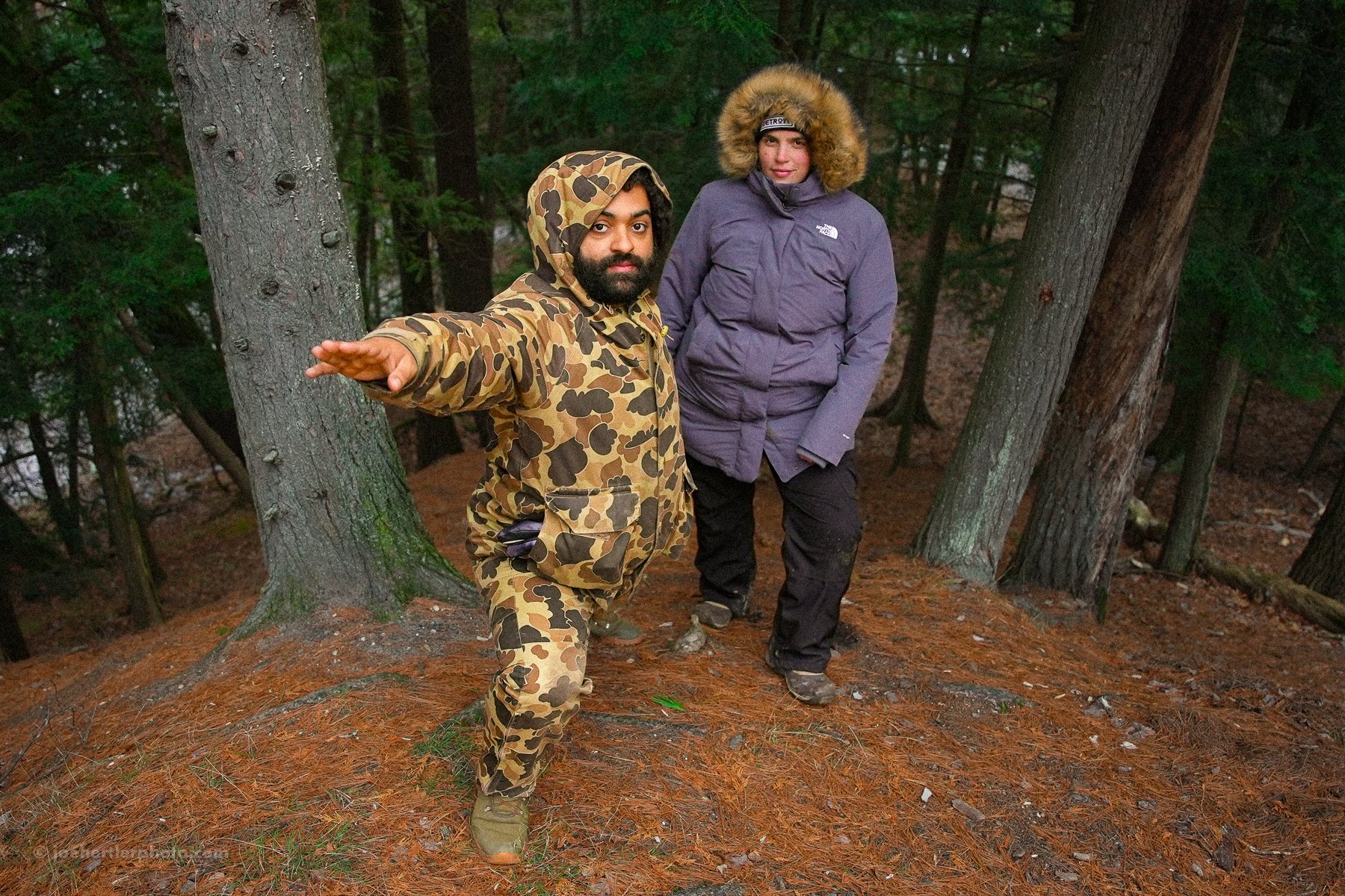 Two people in outdoor winter clothing stand in a wooded forest with tall trees and pine needles on the ground. One person is dressed in a camouflage jacket and pants, with a hood, and is extending an arm. The other person is wearing a purple jacket w