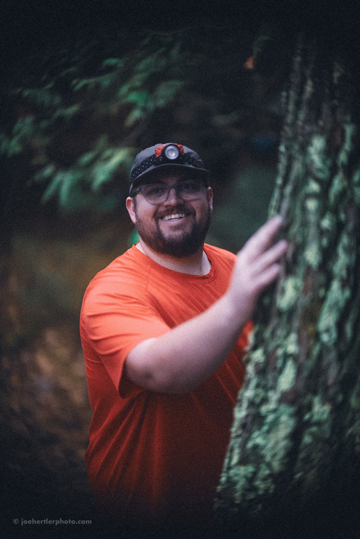 A man in an orange shirt and a headlamp is smiling and standing outdoors next to a tree.
