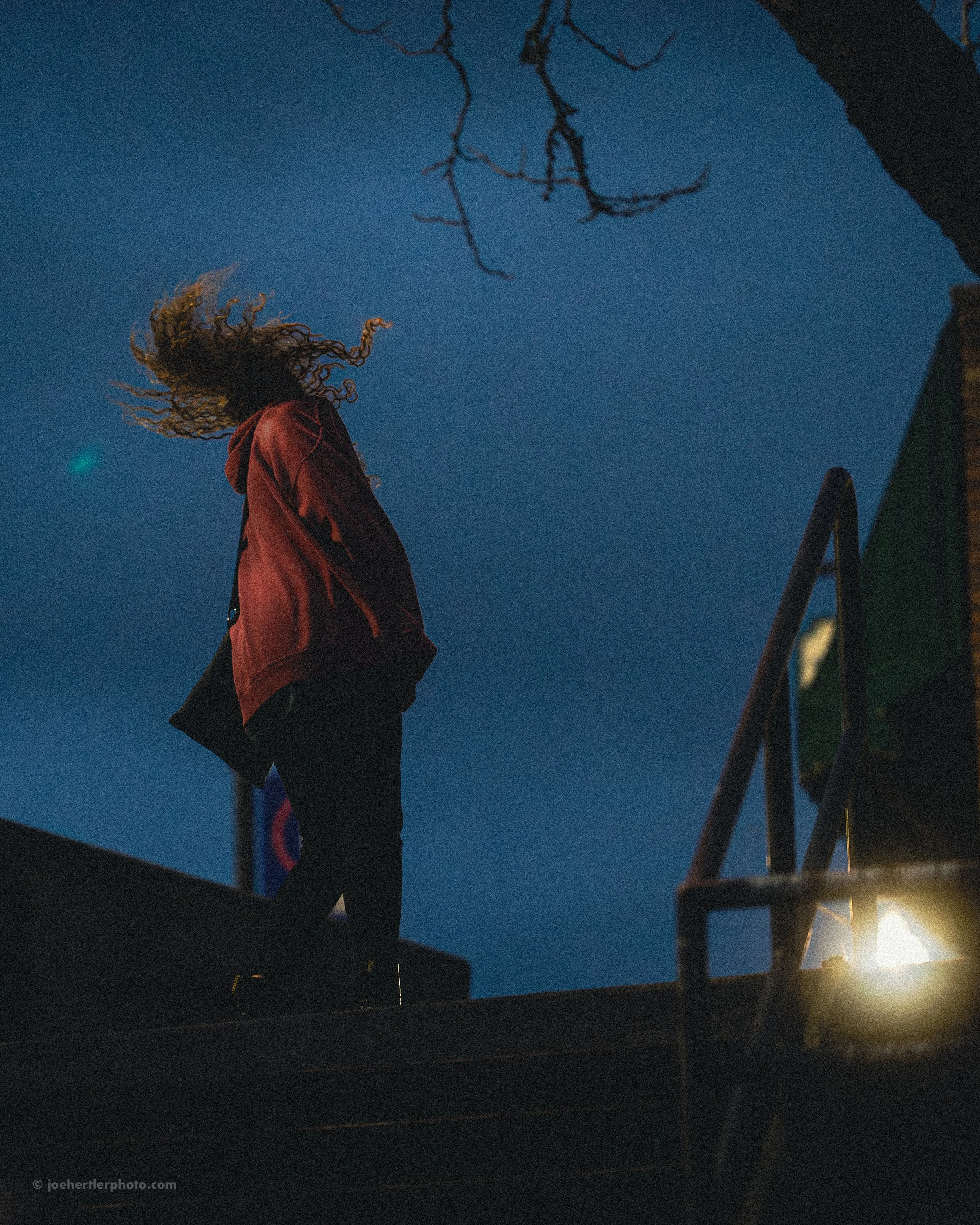 A person with curly hair standing outdoors at dusk or night, wearing a red jacket and black pants, with their head turned away from the camera. The scene features a dark sky with some visible tree branches and a light source illuminating part of a st