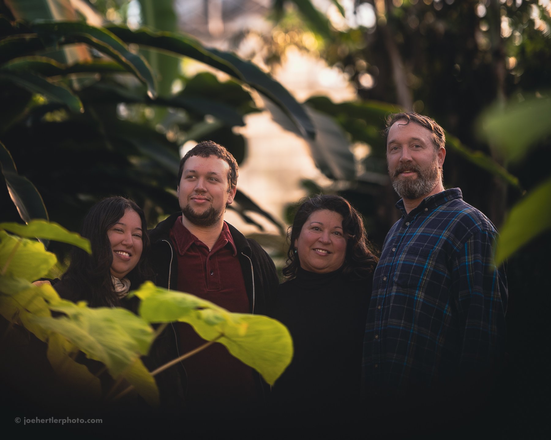 Group of four diverse people standing outdoors amongst large green leaves, smiling at the camera during golden hour.