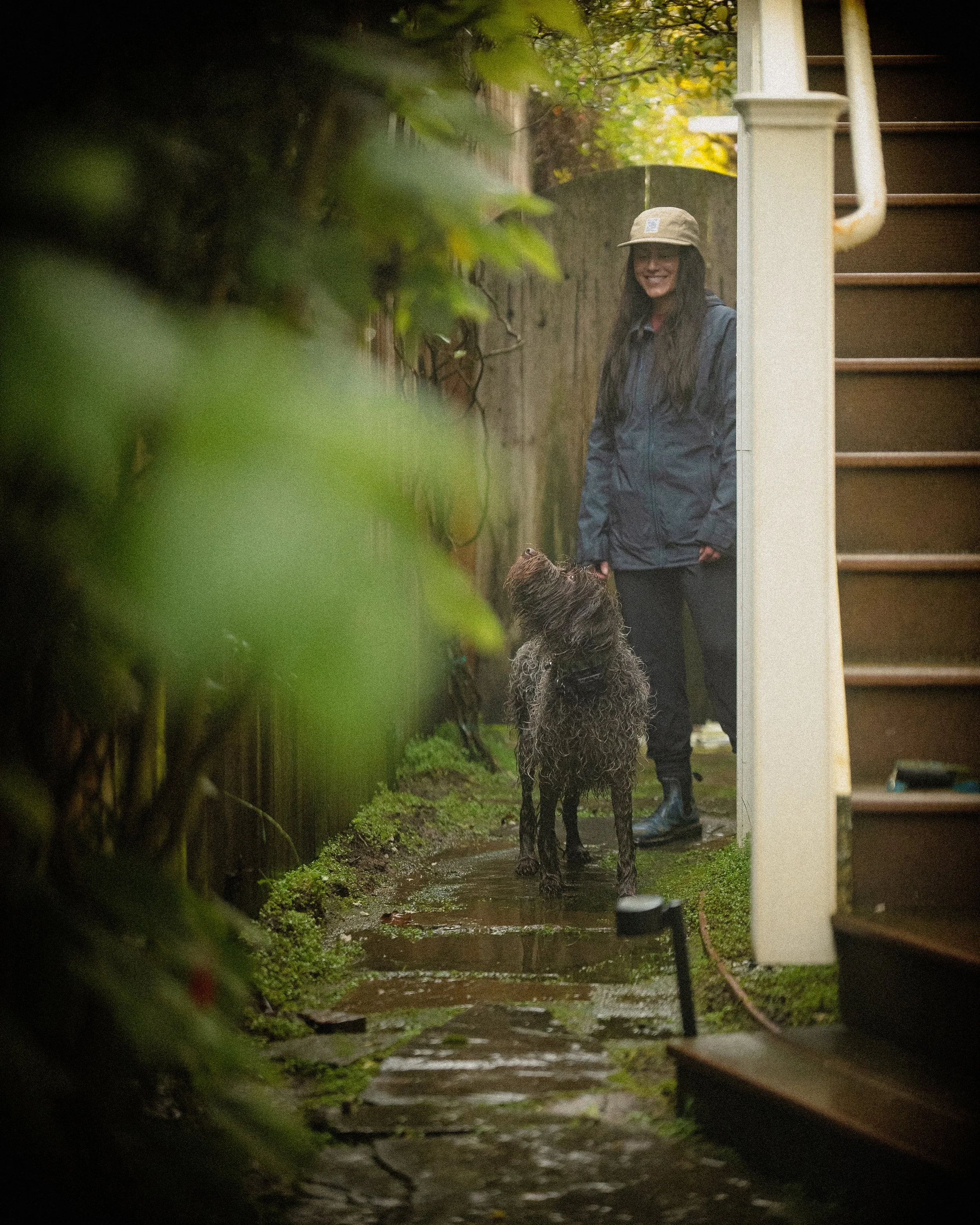 A woman in a blue rain jacket and tan hat stands on a wet pathway next to a staircase, smiling at a wet, curly-haired dog looking up at her.