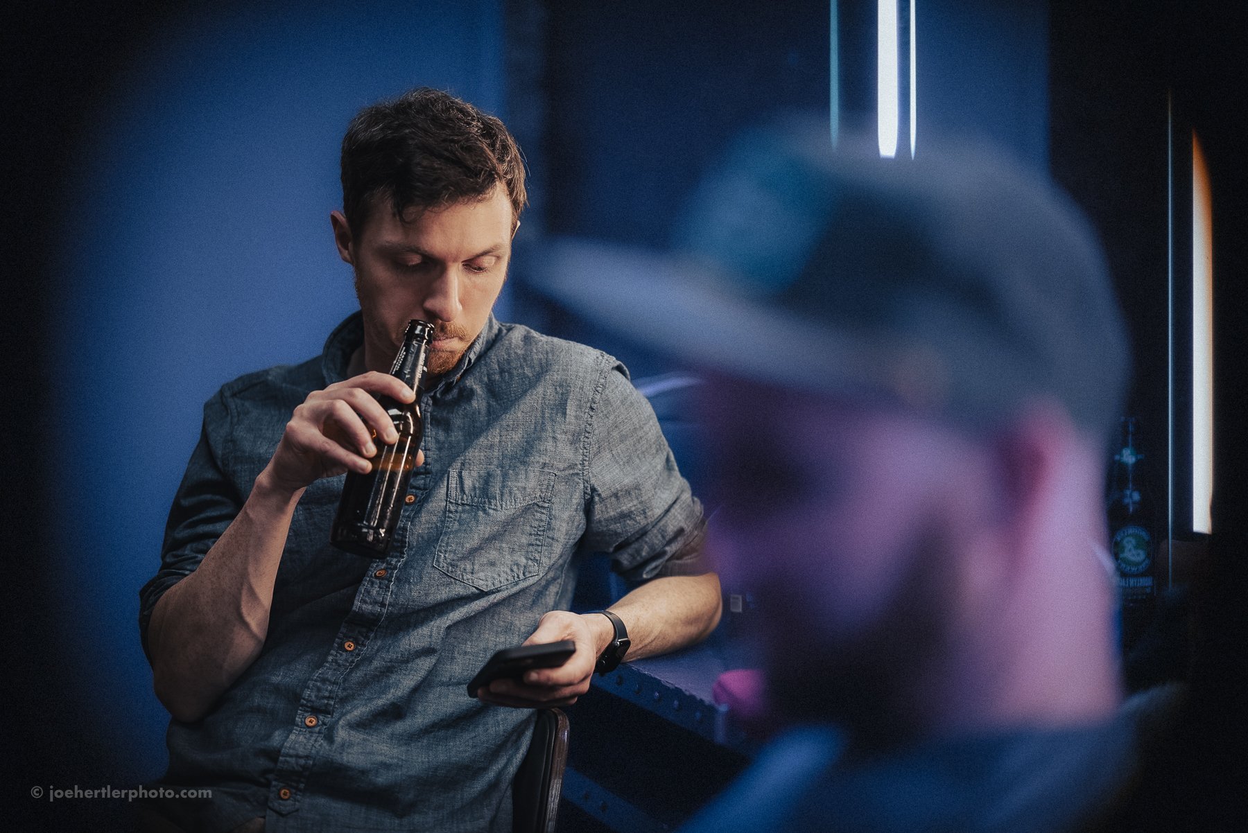 A young man with dark hair and a beard sips from a brown glass bottle while looking at his phone, sitting in a dimly lit room with blue walls and modern lighting in the background.