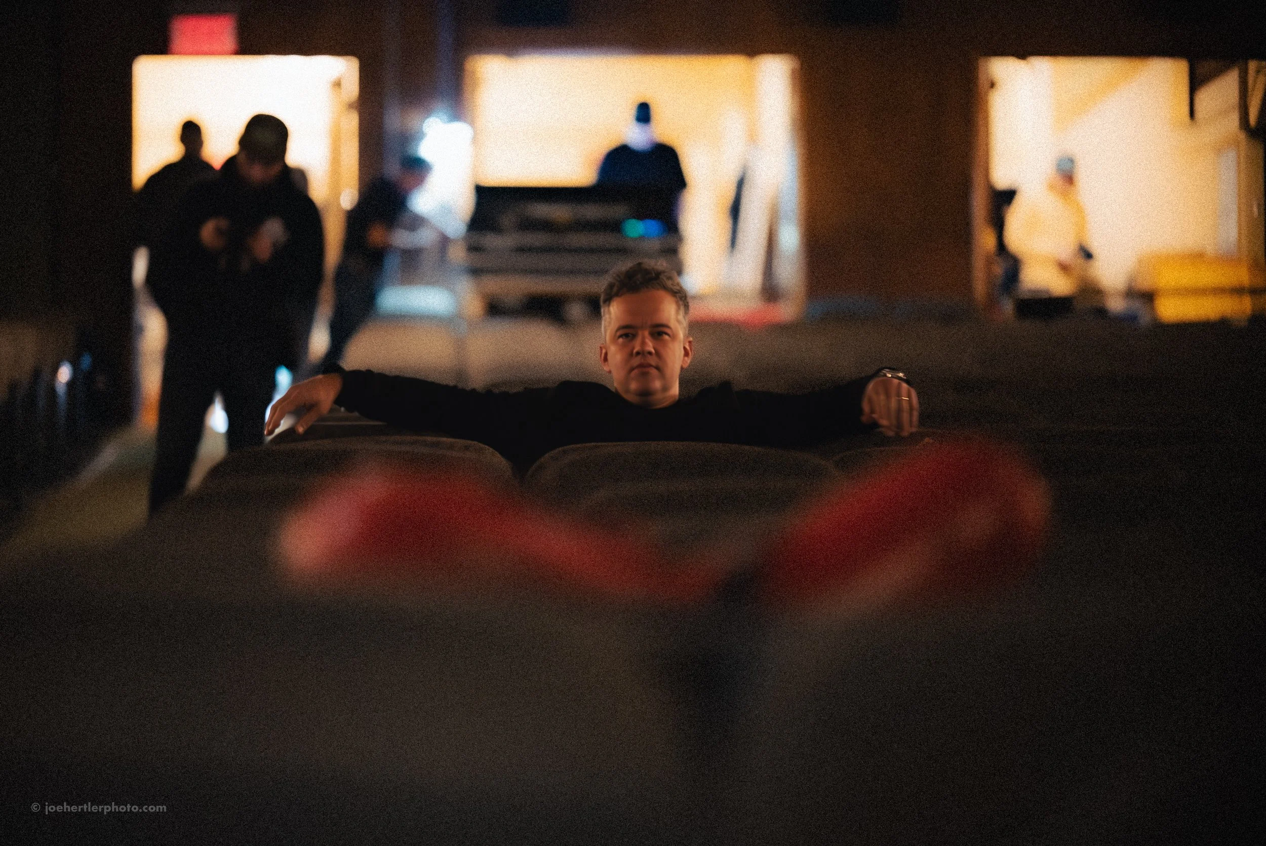 A young man in black clothing sitting with arms stretched out on the backs of theater seats, looking directly at the camera in a dimly lit movie theater. Other people are sitting and standing in the background, with some standing at a counter or behi