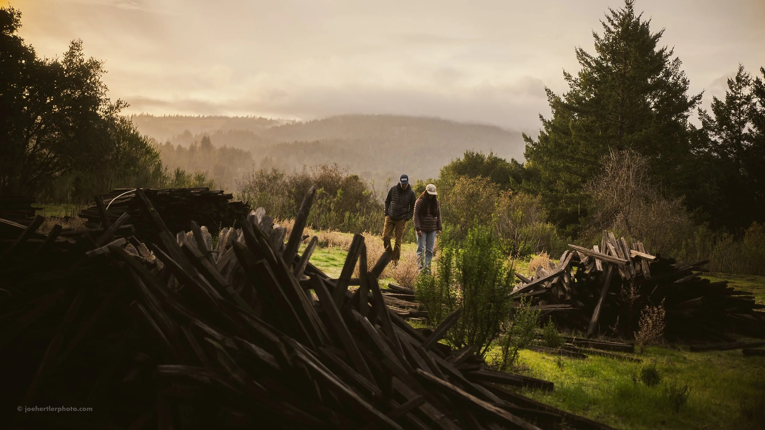A couple walks along a grassy trail surrounded by piles of dark wooden planks and lush green trees, with a mountain range in the distance under a cloudy sky.