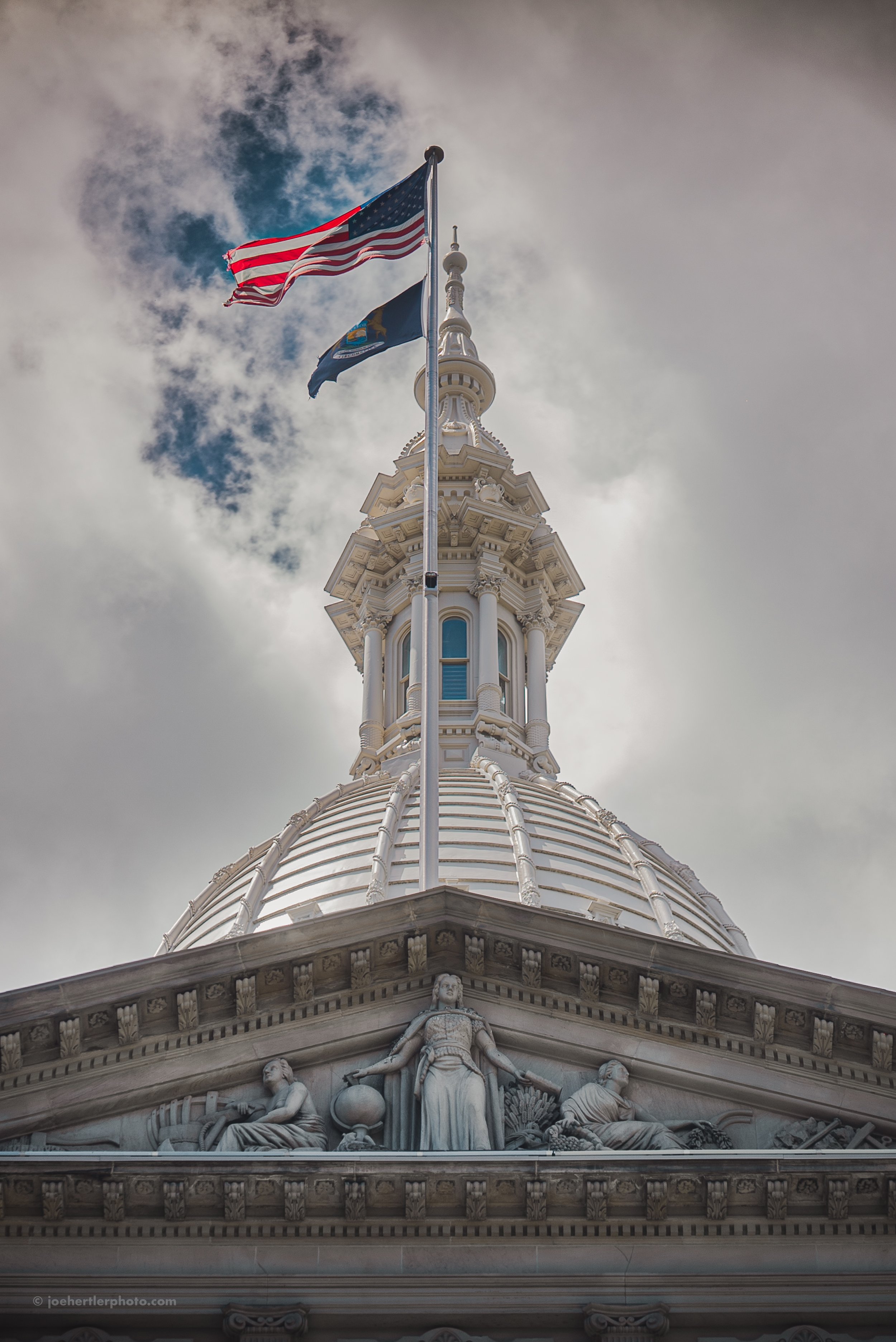 Close-up of a domed architectural building with sculptures and an American flag and a state flag flying above, against a cloudy sky.