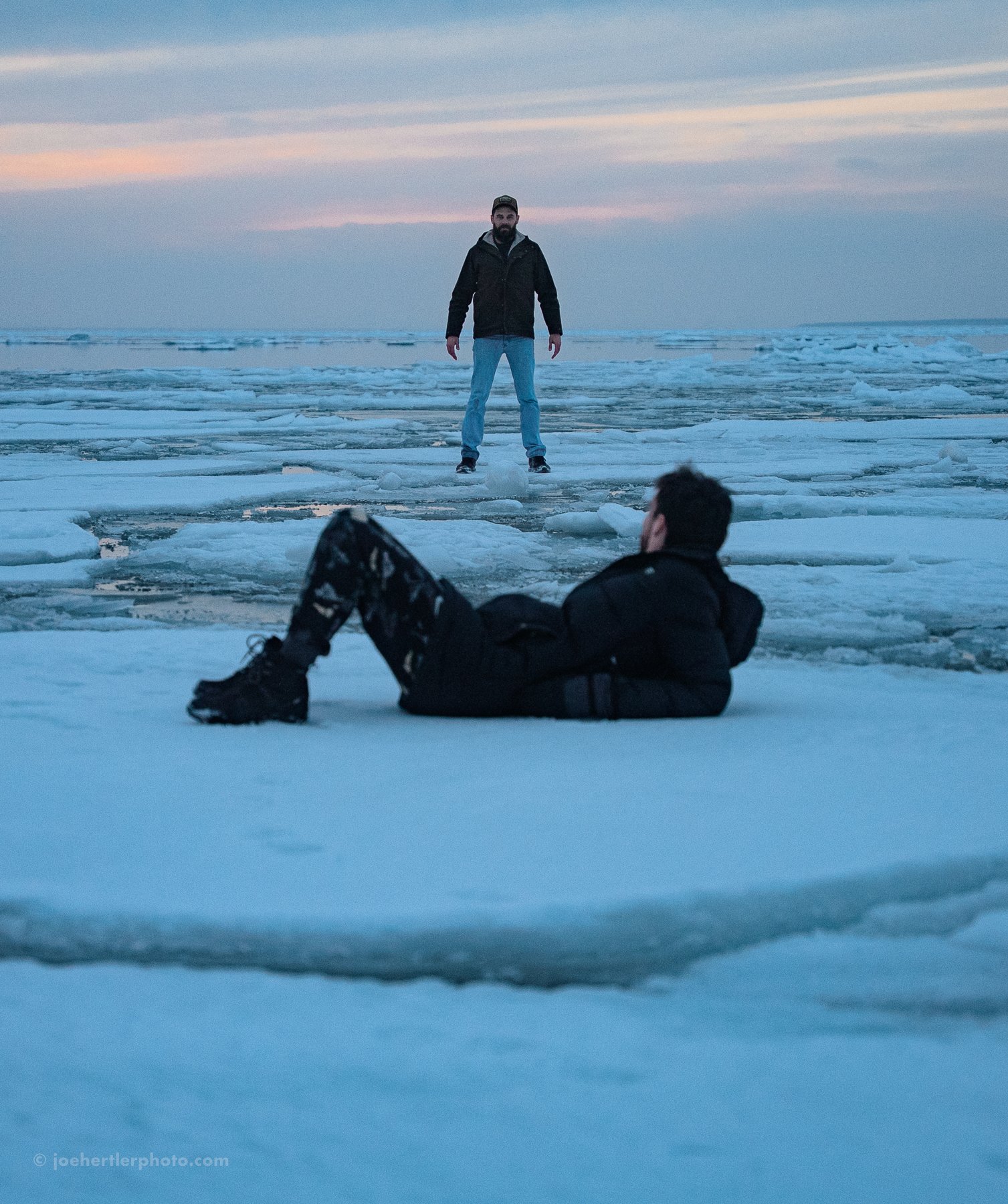 Two men in winter clothing on a frozen icy landscape, one standing and one lying down, with a sunset sky in the background.