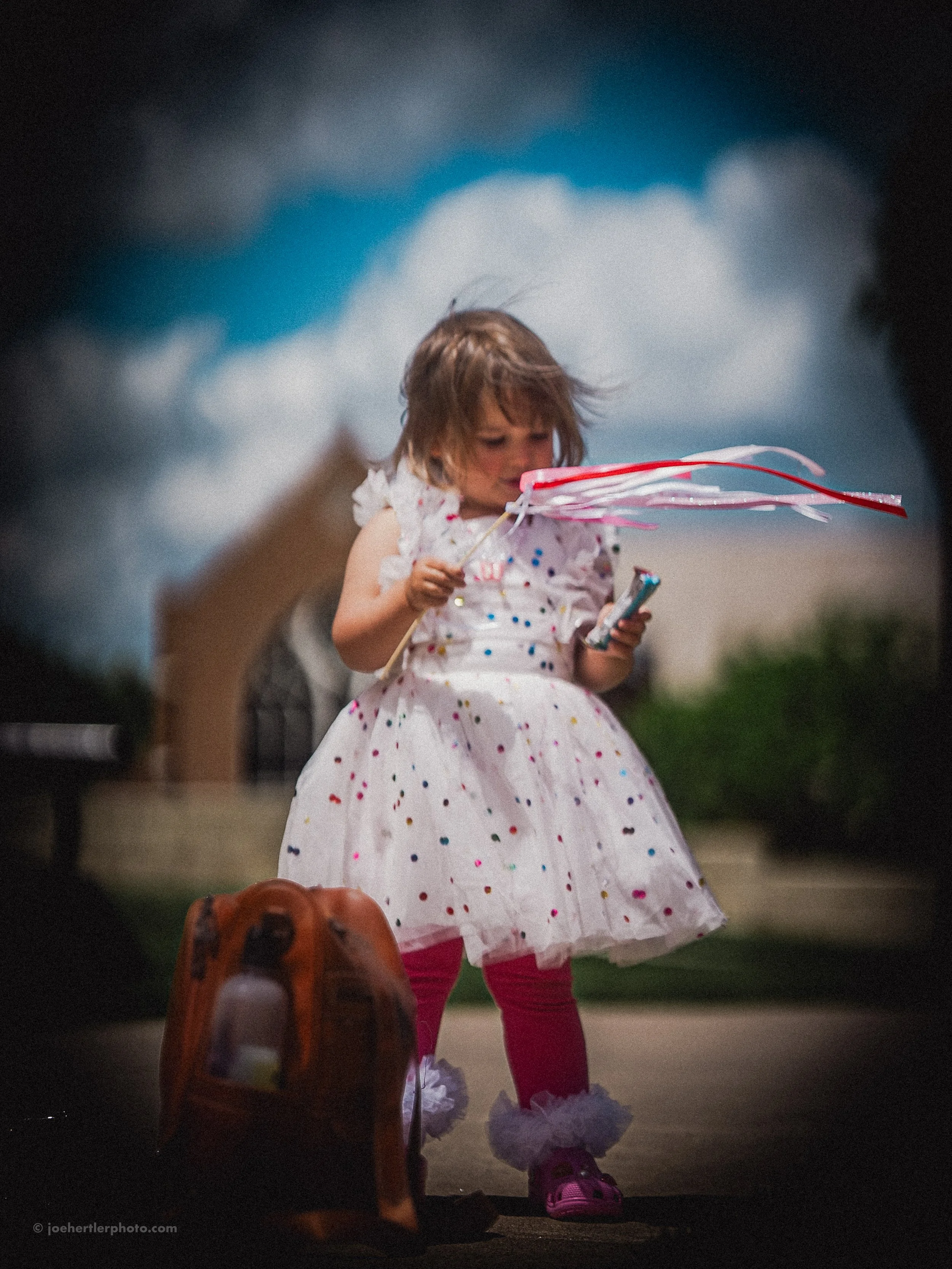 A young girl in a white dress with colorful polka dots and pink leggings, holding a pink and white kite, stands outside with a cloudy sky and a building in the background. A backpack is on the ground nearby.