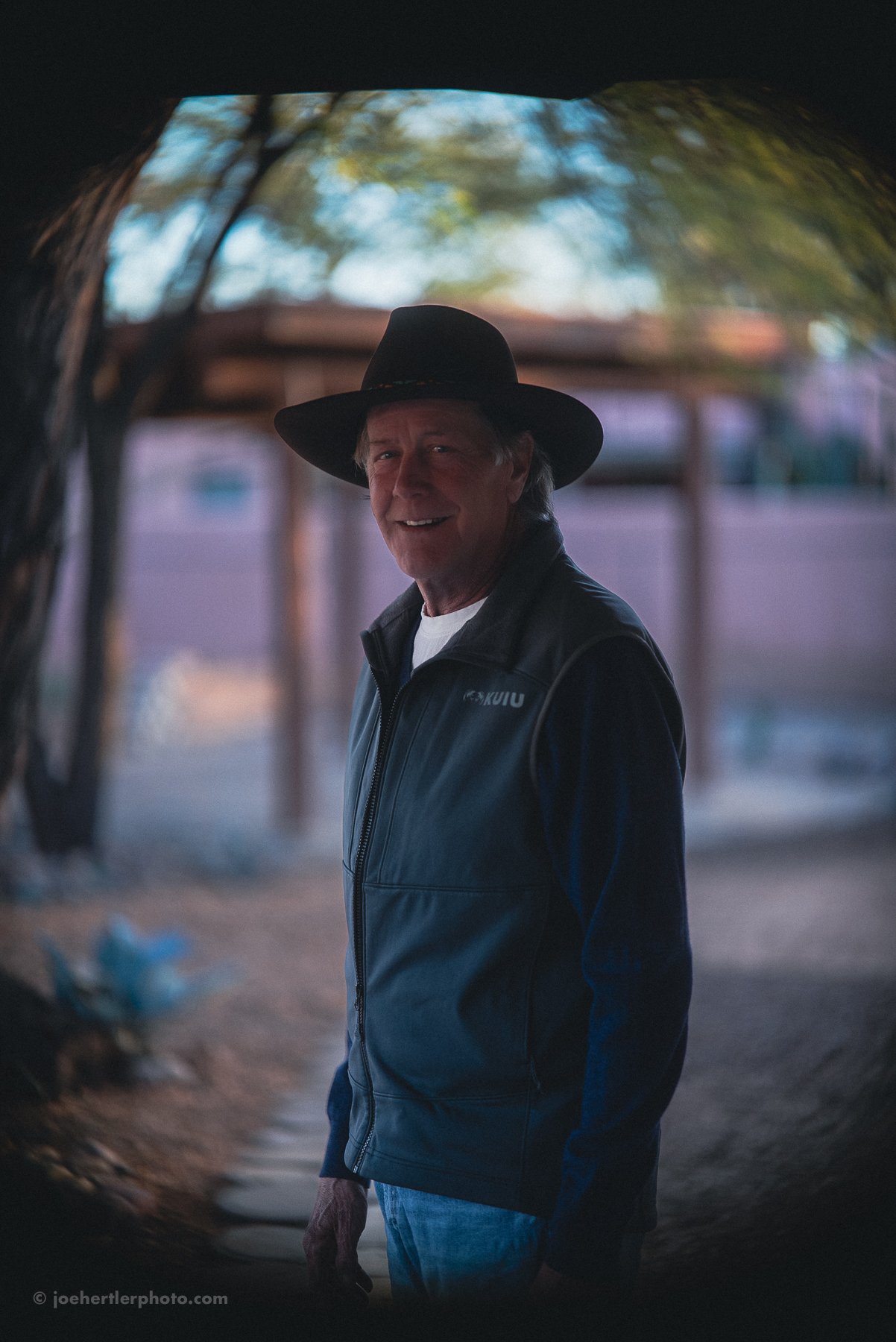 An older man wearing a black wide-brimmed hat and a dark jacket, smiling and looking toward the camera, standing outdoors in a shaded area with a blurred background of trees and structures.