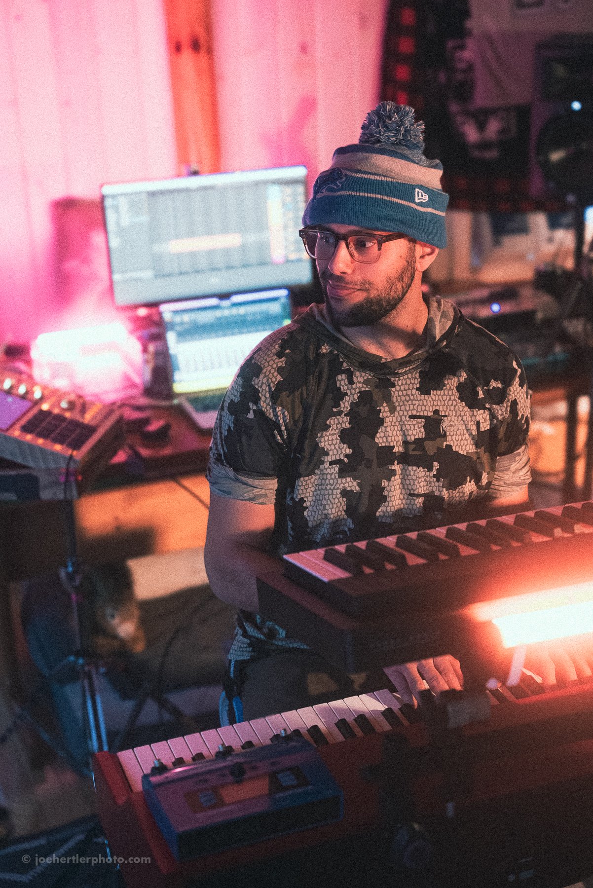 A man in a patterned shirt, wearing a beanie, glasses, and headphones, sits at a keyboard in a music studio with electronic equipment and monitors in the background, illuminated by pink and purple lighting.
