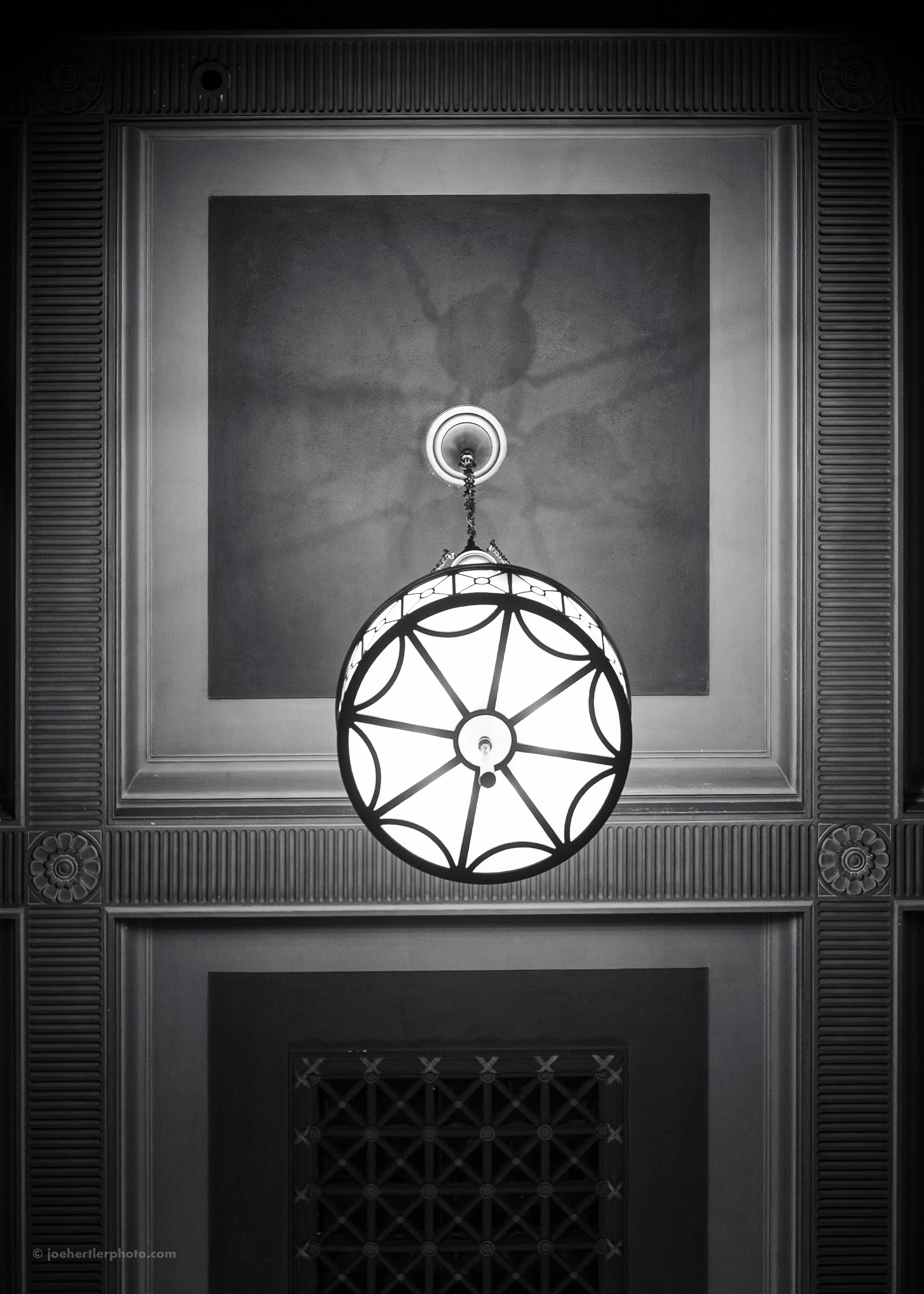 Black and white photo of a ceiling light fixture casting a shadow that looks like a face with outstretched arms, mounted on a decorative ceiling with ornate molding and a square ceiling panel.