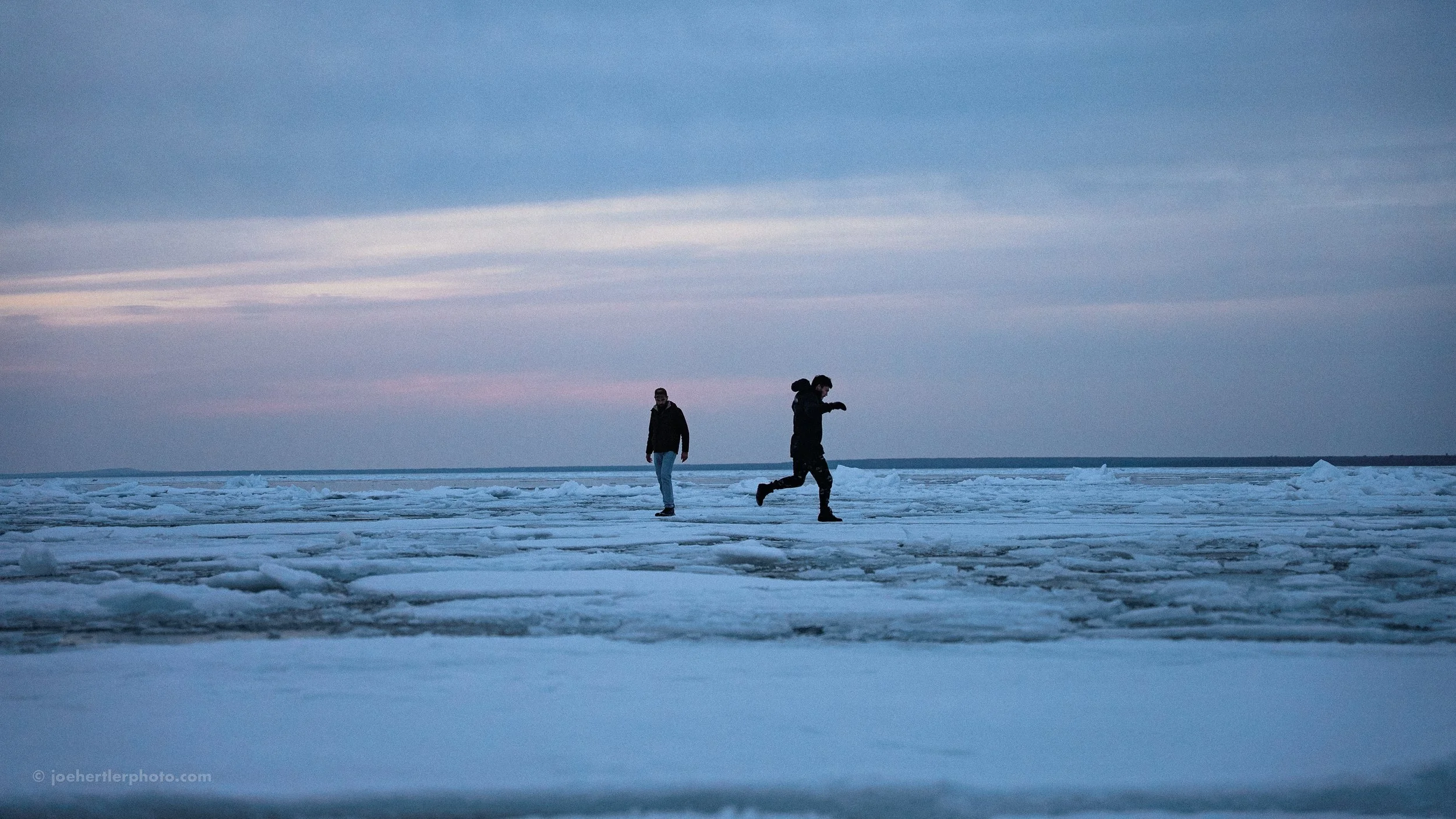 Two people walking on a frozen icy landscape, with one person running and the other walking, under a cloudy sky during sunset or sunrise.