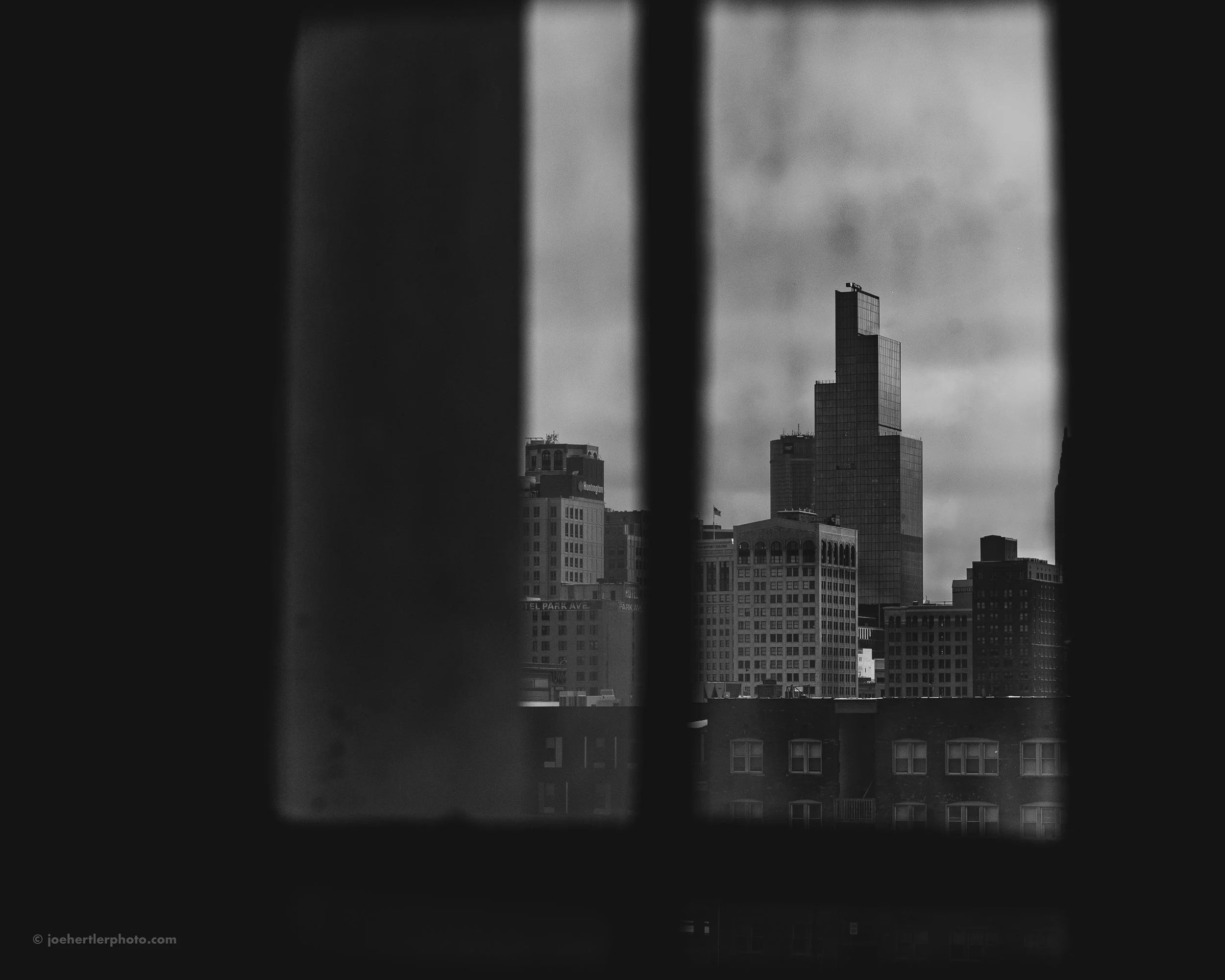 Black and white photograph of city skyline viewed through a barred window with vertical bars, featuring tall modern skyscrapers.