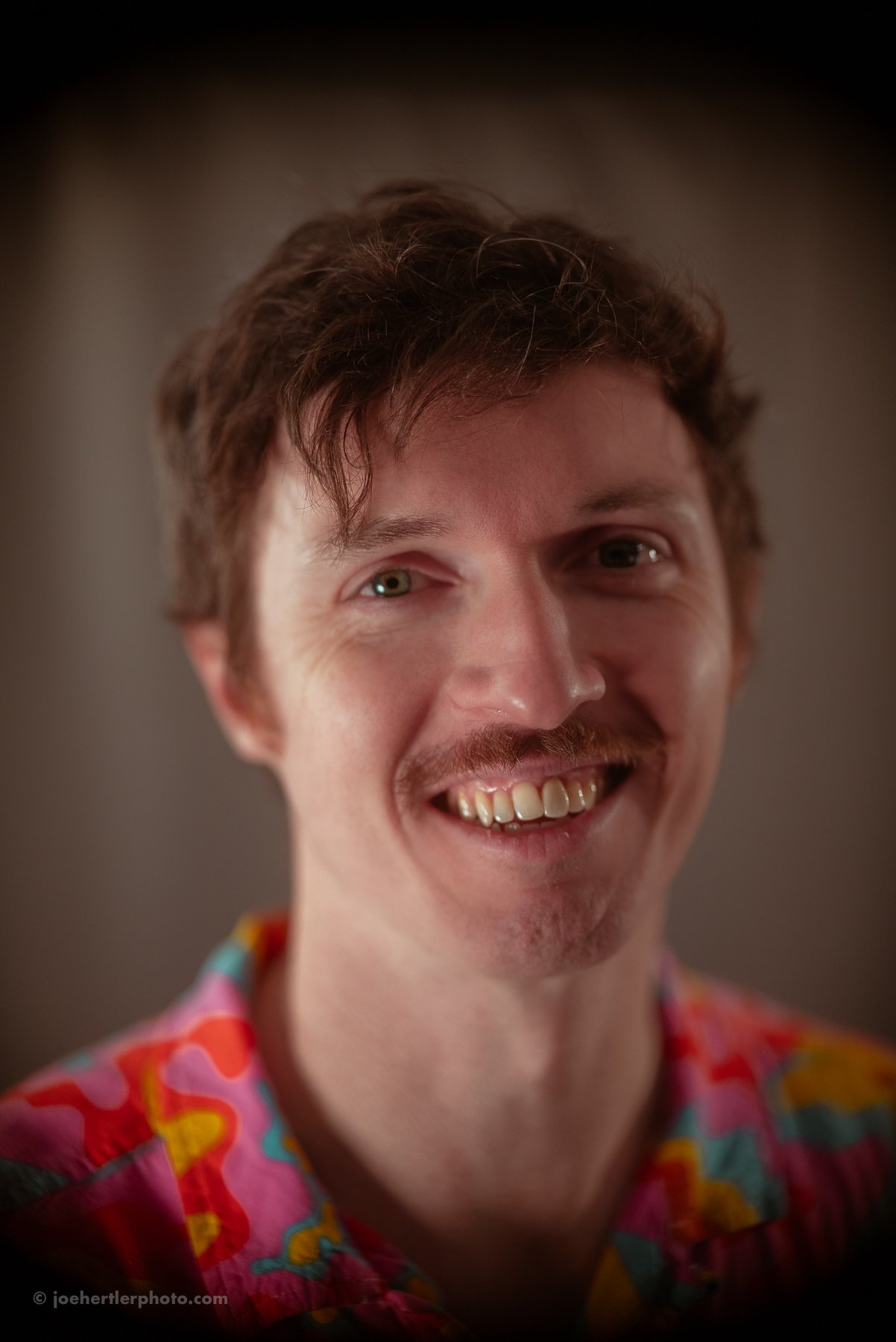 Close-up of a man with curly brown hair, green eyes, a mustache and goatee, smiling, wearing a colorful, patterned shirt.