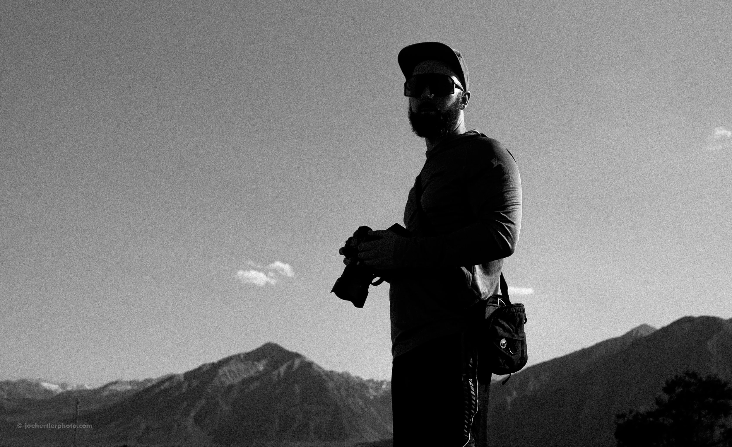 A silhouette of a man standing outdoors, holding an object in his hands, with mountain ranges in the background on a clear day.