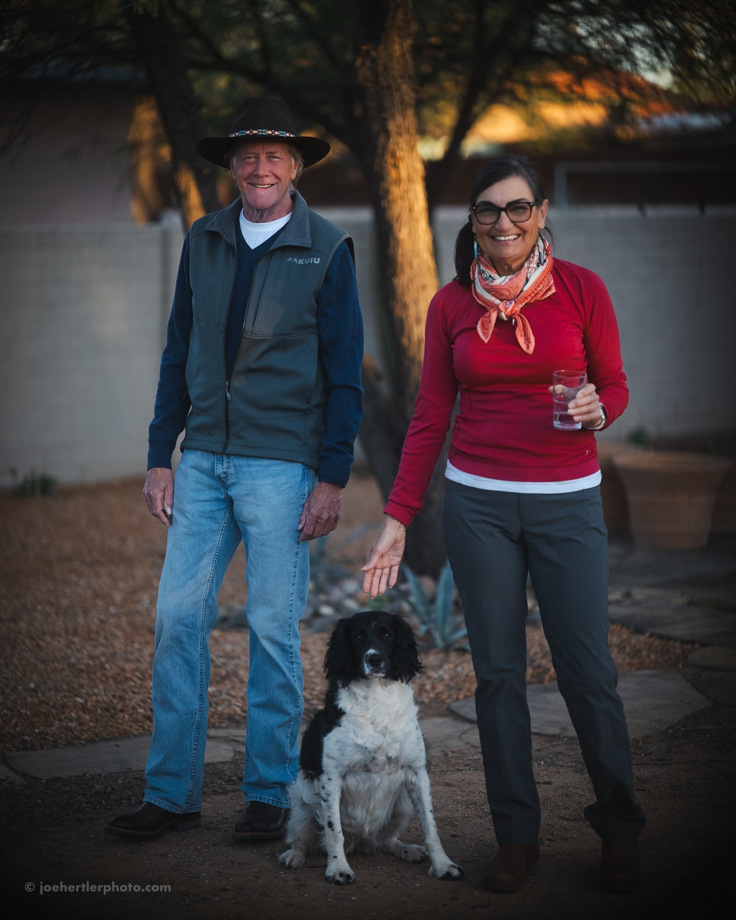 A man and a woman standing outside with a black and white dog, smiling. The man is wearing a hat, a green vest, and jeans, and the woman is holding a glass of water, wearing glasses, a red shirt, and a scarf. The background shows trees and a fence at