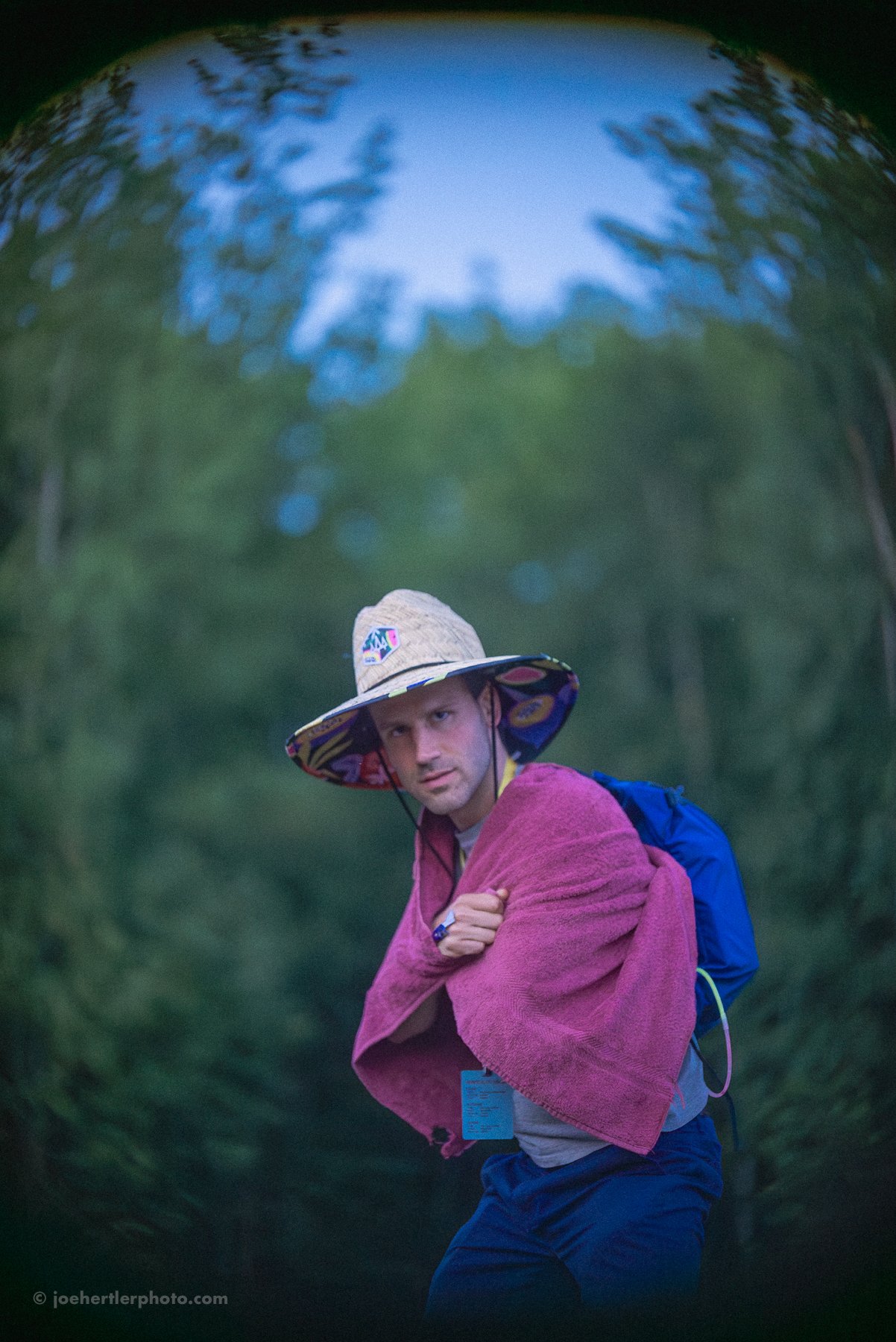 A man wearing a wide-brimmed hat with patterns, a pink towel draped over his shoulders, a gray shirt, and carrying a blue backpack, standing outdoors with a background of green trees and sky