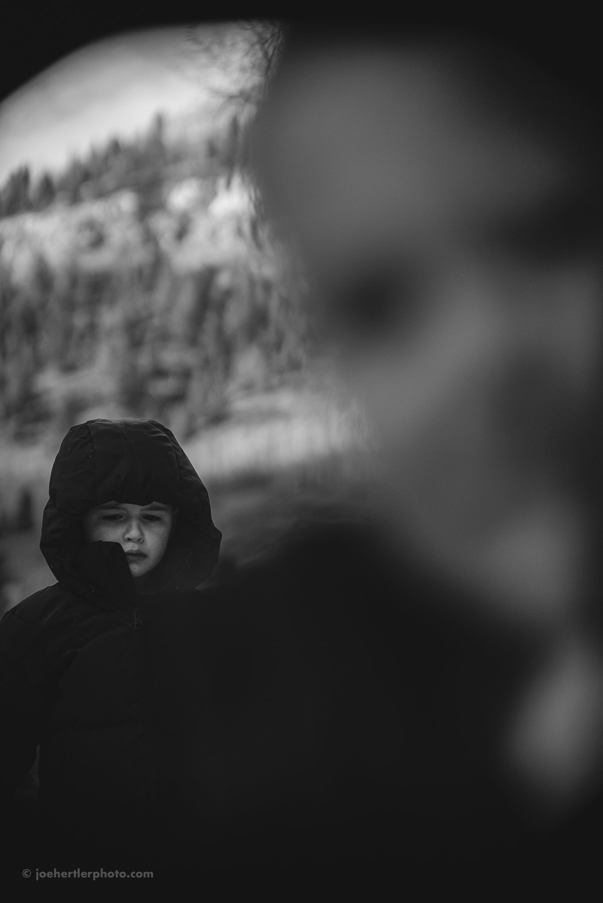 A young child wearing a hooded jacket, standing outdoors during what appears to be cold weather, with trees blurred in the background.