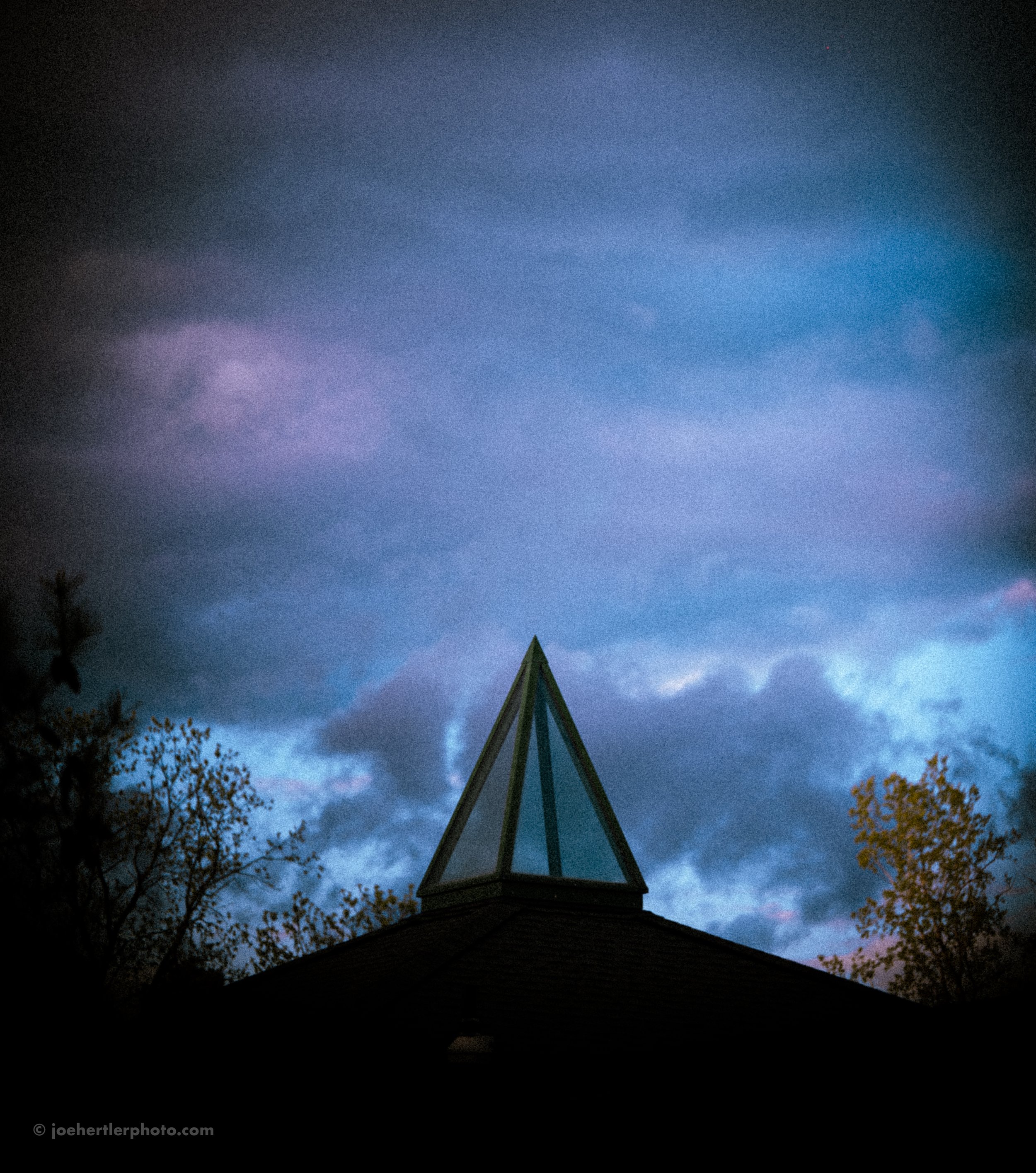 A building with a glass pyramid-shaped window on its roof against a cloudy evening sky with trees on both sides.